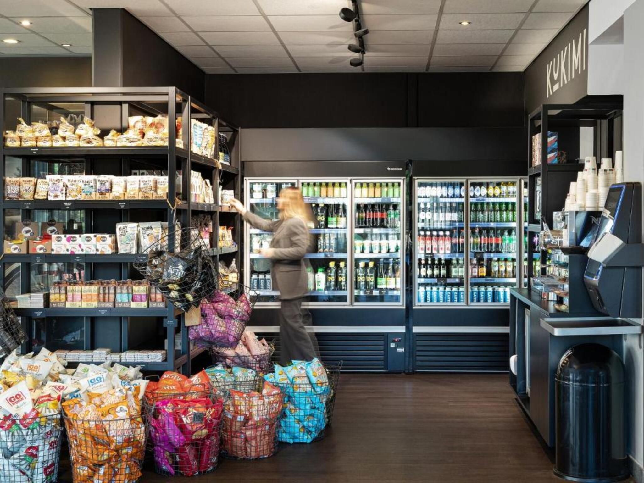 A grocery store interior with shelves stocked with snacks, a refrigerated section with beverages, and a checkout area with a self-service machine.