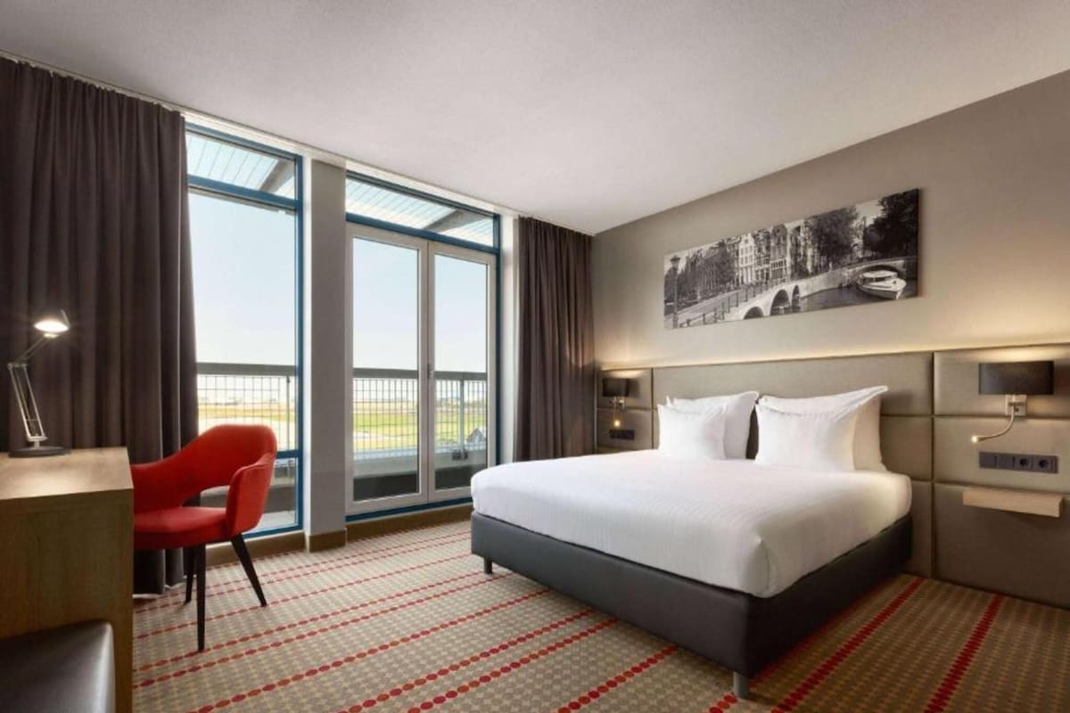 Habitación de hotel con una cama grande, un escritorio, una silla y vistas al exterior a través de las ventanas.