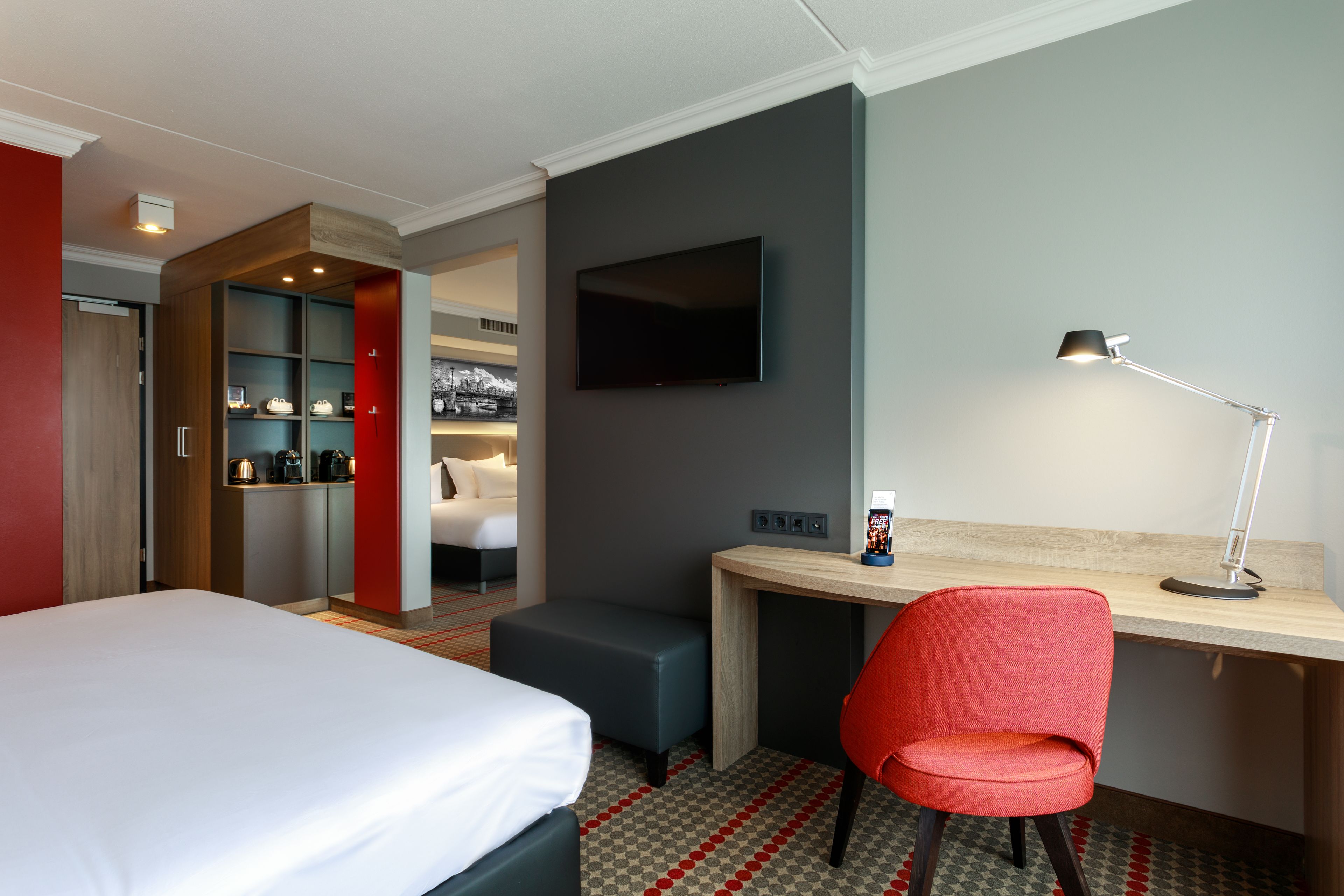 A hotel room with a bed, a desk, a red chair, a TV, and a coffee machine.