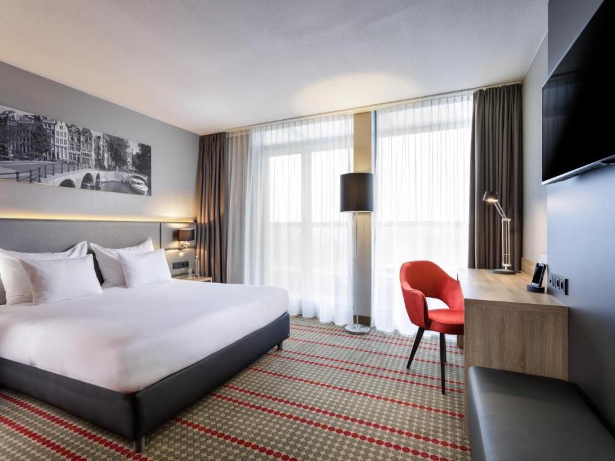 A hotel room with a large bed, a desk, a chair, a TV, and a window with curtains.