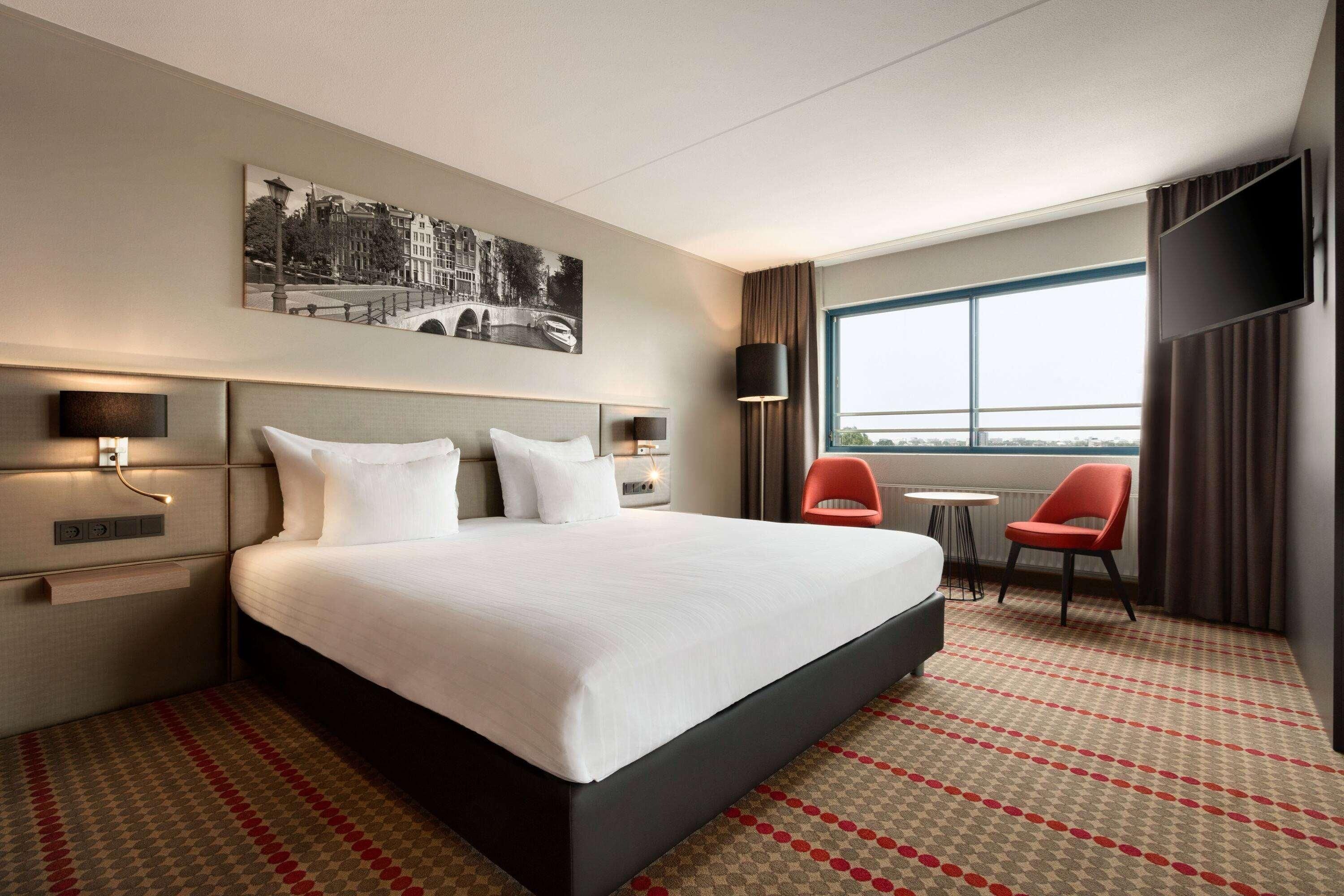 Una habitación de hotel con una cama grande, una mesita pequeña, dos sillas, un televisor y una ventana con vistas a la ciudad.