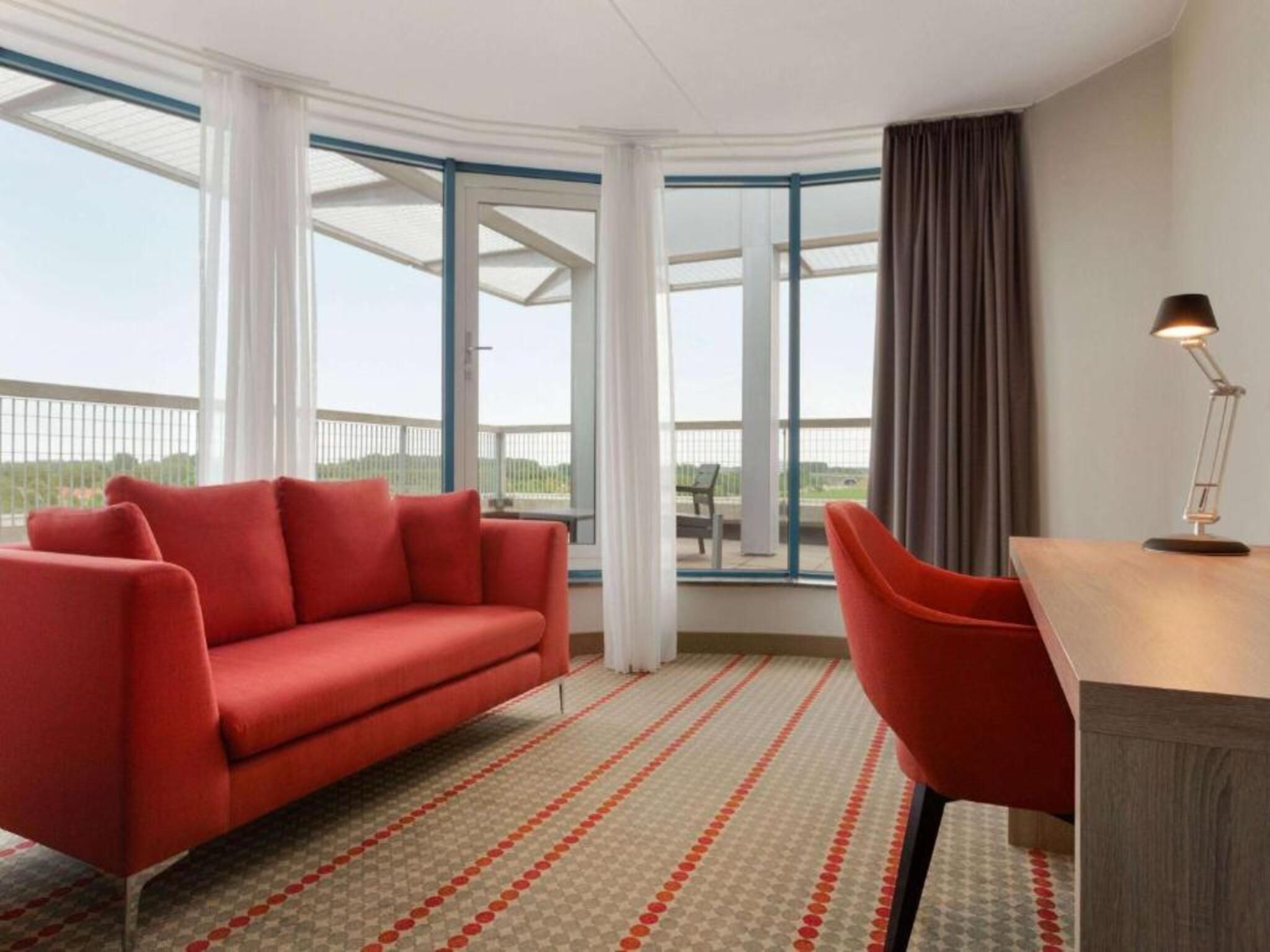 Una habitación moderna con un sofá rojo, un escritorio y grandes ventanales que ofrecen vistas al exterior.