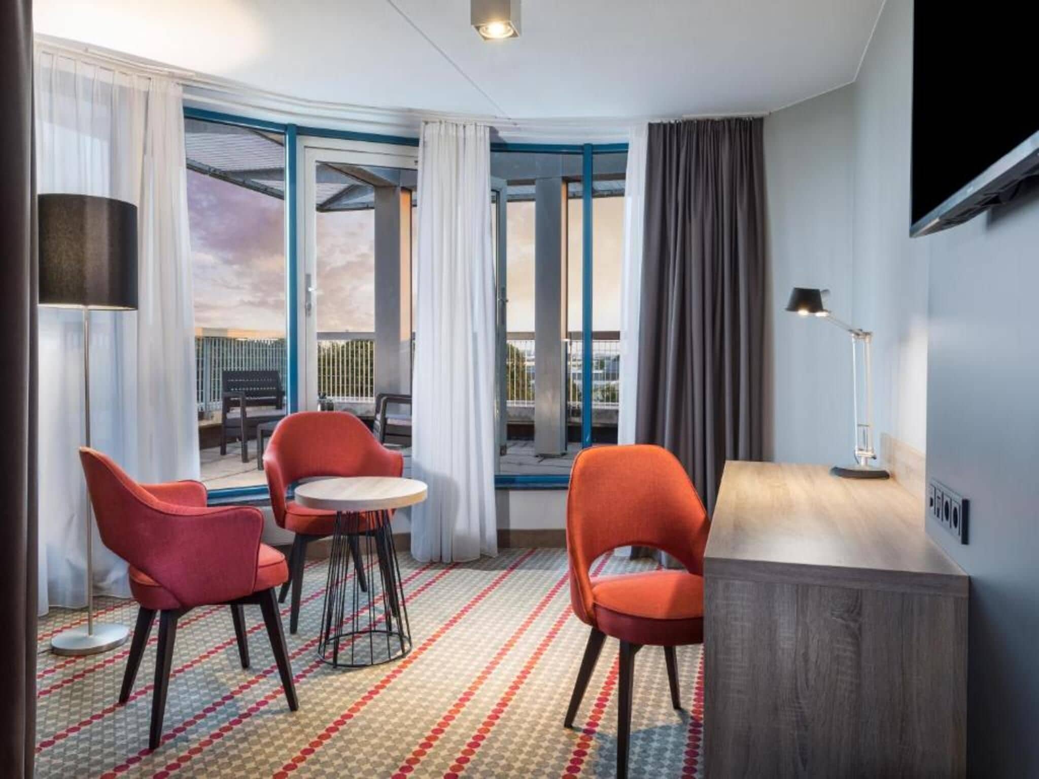 Ein Hotelzimmer mit einem Bett, zwei roten Sesseln, einem kleinen Tisch, einem Fernseher und Blick ins Freie.