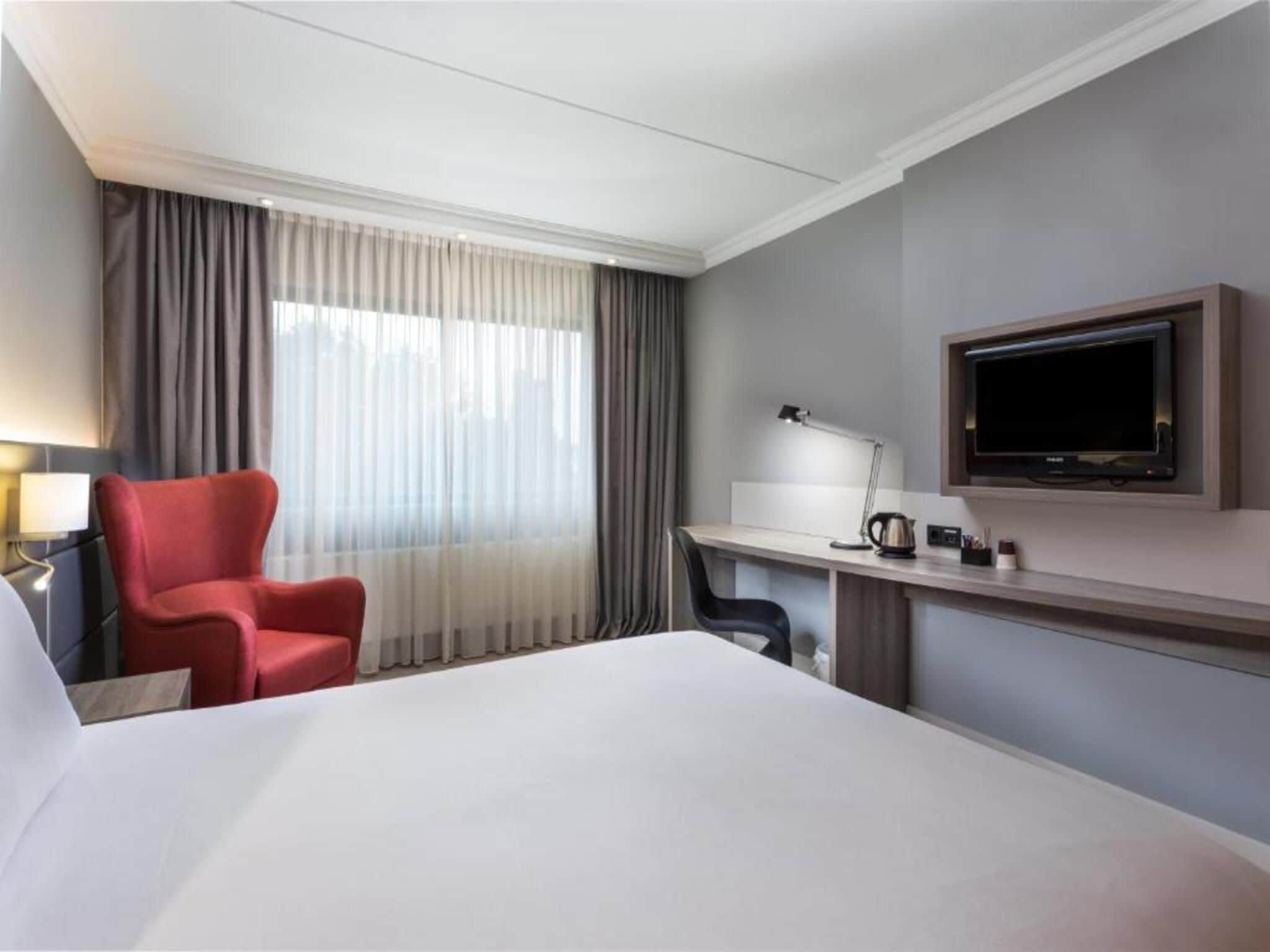 Habitación de hotel con cama, escritorio, silla, televisor y cortinas.
