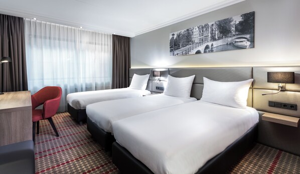 A hotel room with two beds, a desk, a chair, and a large window with sheer curtains.
