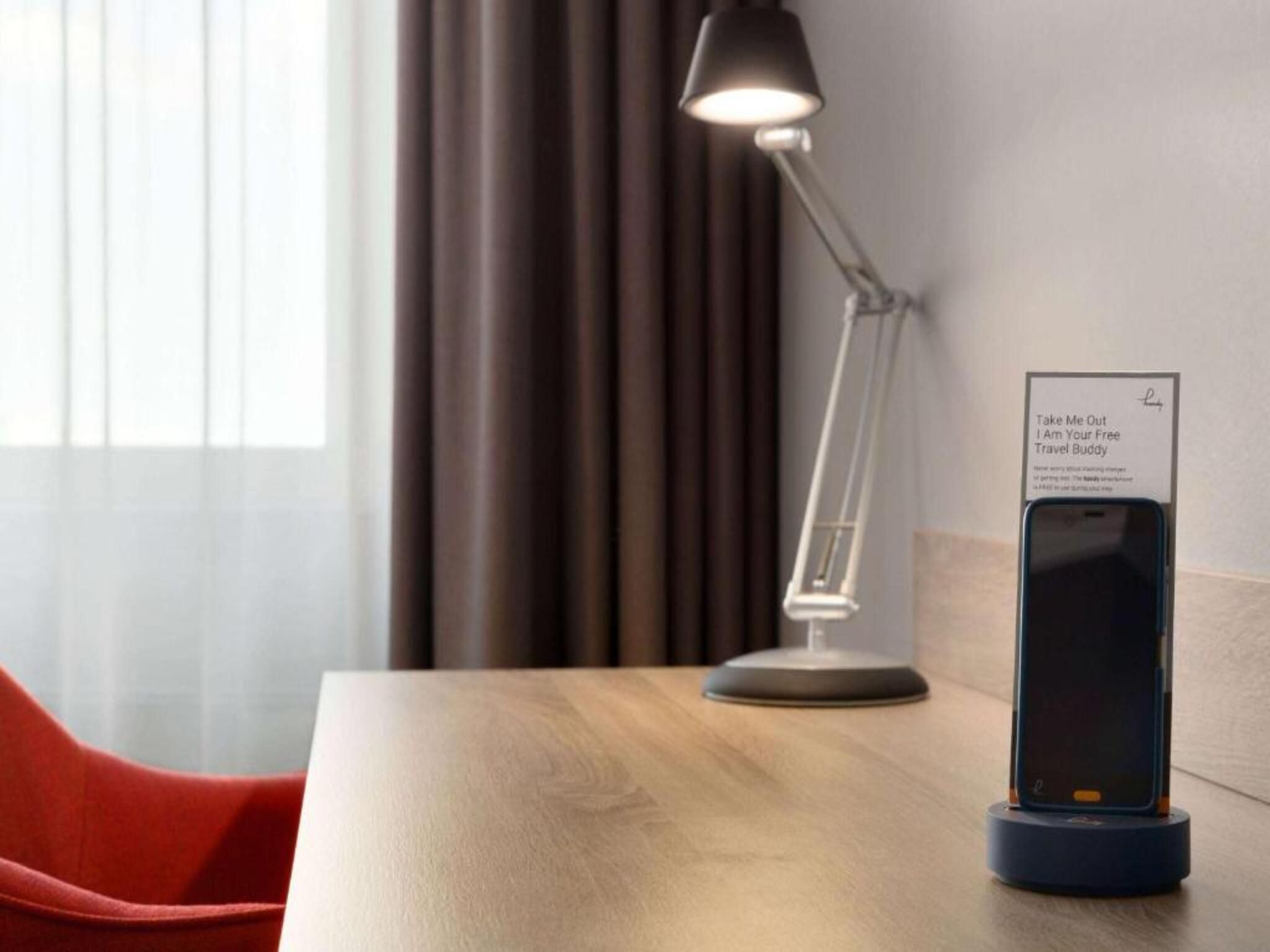 Habitación de hotel con escritorio, lámpara de escritorio moderna, soporte para cargar un teléfono inteligente y un cartel que ofrece servicio de acompañante de viaje gratuito.