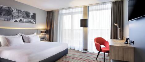 Ein Hotelzimmer mit einem großen Bett, einem Schreibtisch mit Stuhl, einem Flachbildfernseher und einem großen Fenster mit Vorhängen.