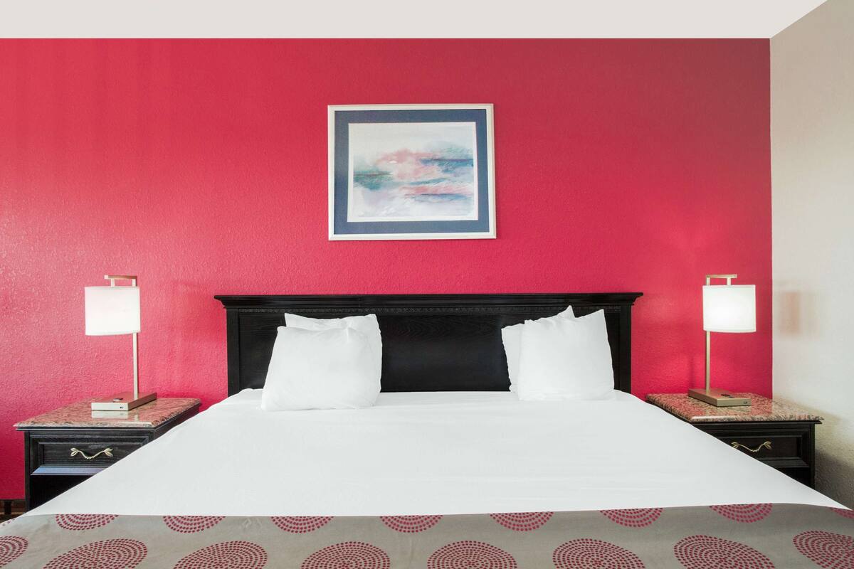 A hotel room with a large bed, two bedside tables, a nightstand, a framed picture on the wall, and a red accent wall.
