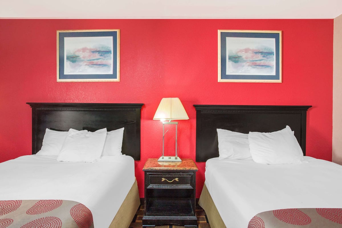 A hotel room with two beds, a nightstand with a lamp, and two framed pictures on the red wall.