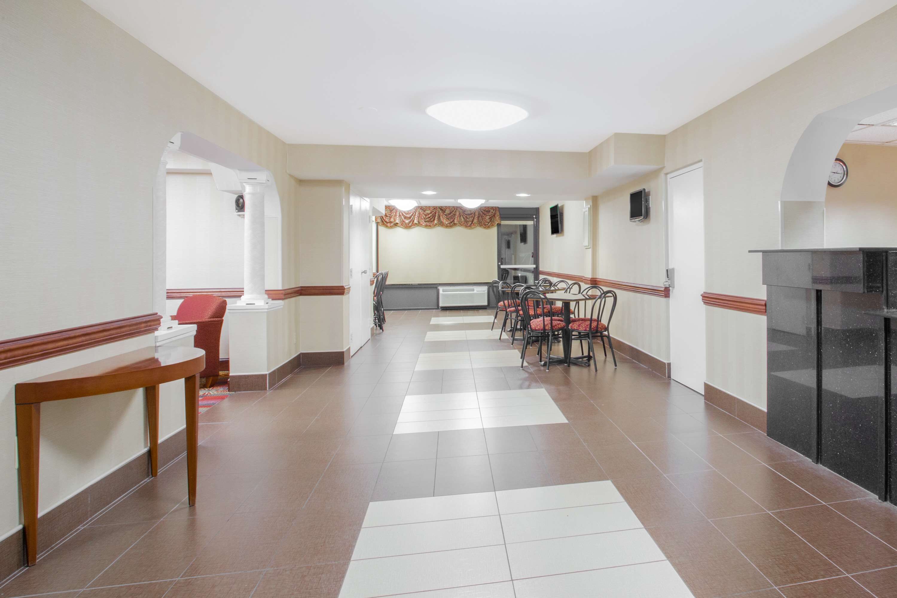 A hotel corridor with a reception desk, a dining area with chairs and tables, and a kitchen area.