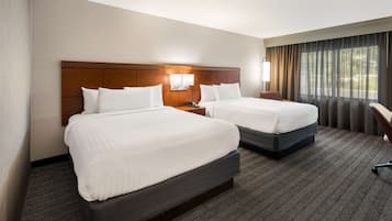 A hotel room with two beds, a wooden headboard, a nightstand with a lamp, and a view of trees through the window.