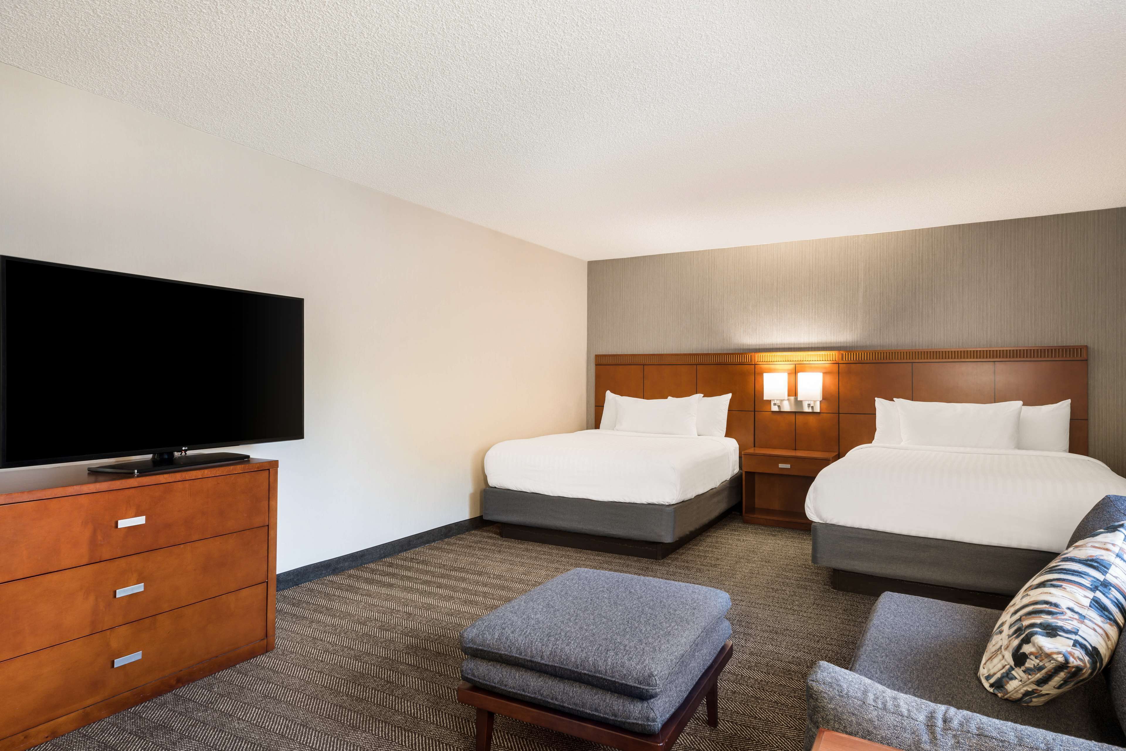 A hotel room with two beds, a television, a wooden dresser, and a small ottoman.