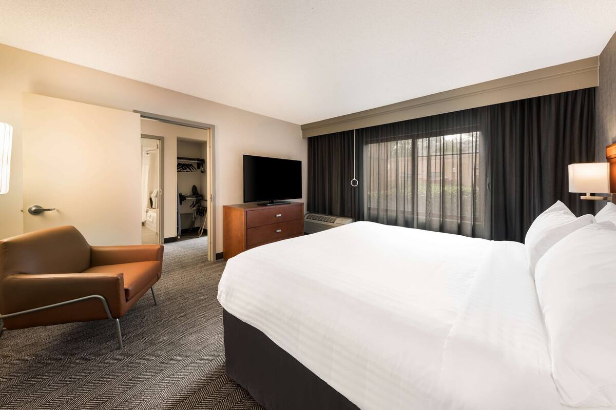 Habitación de hotel con una cama grande, un sillón de cuero marrón, un aparador de madera, un televisor de pantalla plana y un ventanal con cortinas.