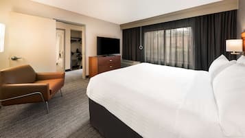 A hotel room with a large bed, a brown leather chair, a wooden dresser, a flat-screen TV, and a large window with curtains.