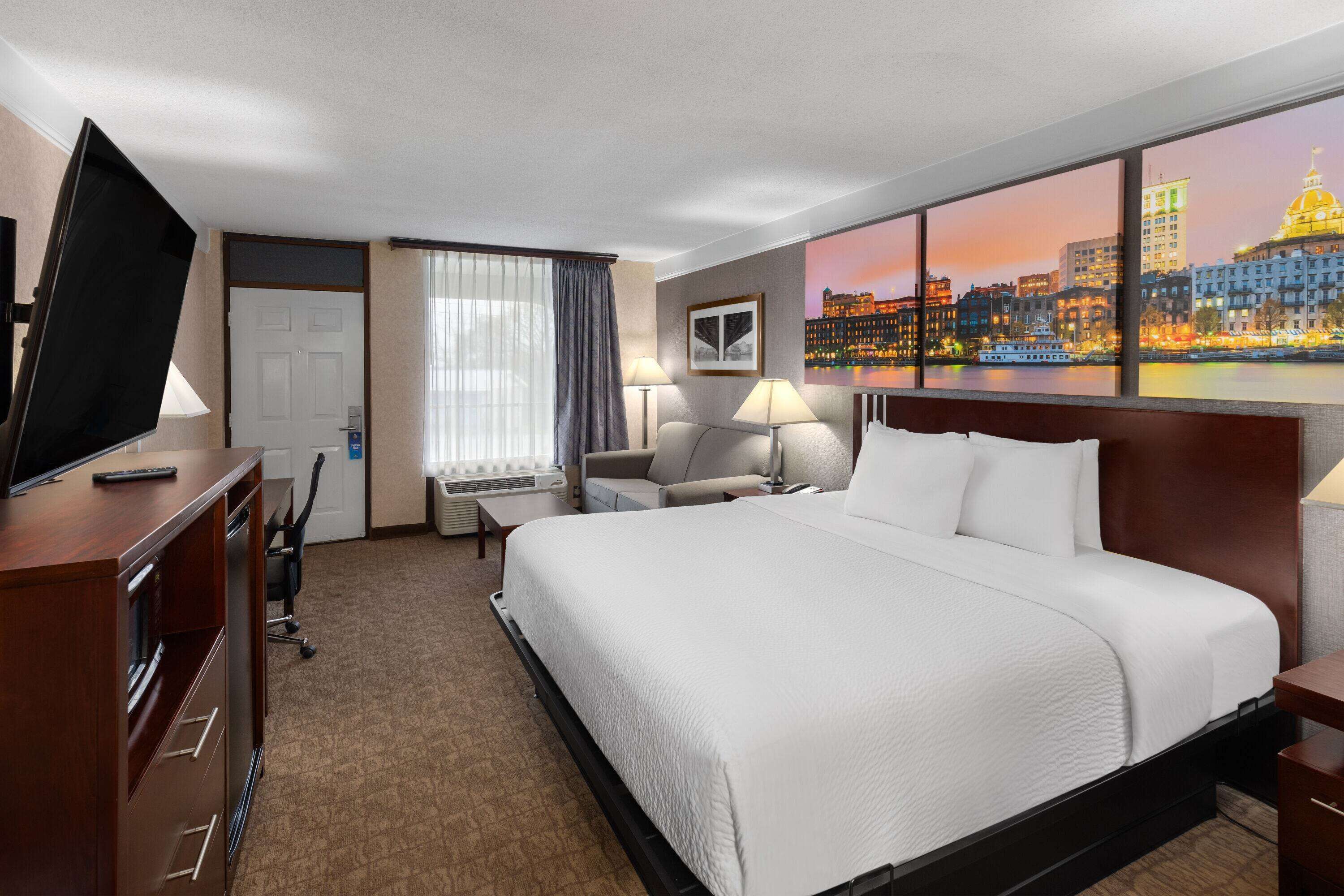 Habitación de hotel con una cama grande, un escritorio con televisor, una silla, una lámpara y un cuadro de un paisaje urbano.