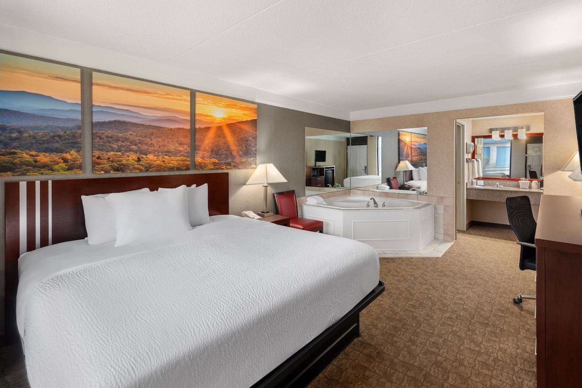 Hotelkamer met een groot bed, een badkamer met bad, een bureau en uitzicht op een zonsondergang door grote ramen.