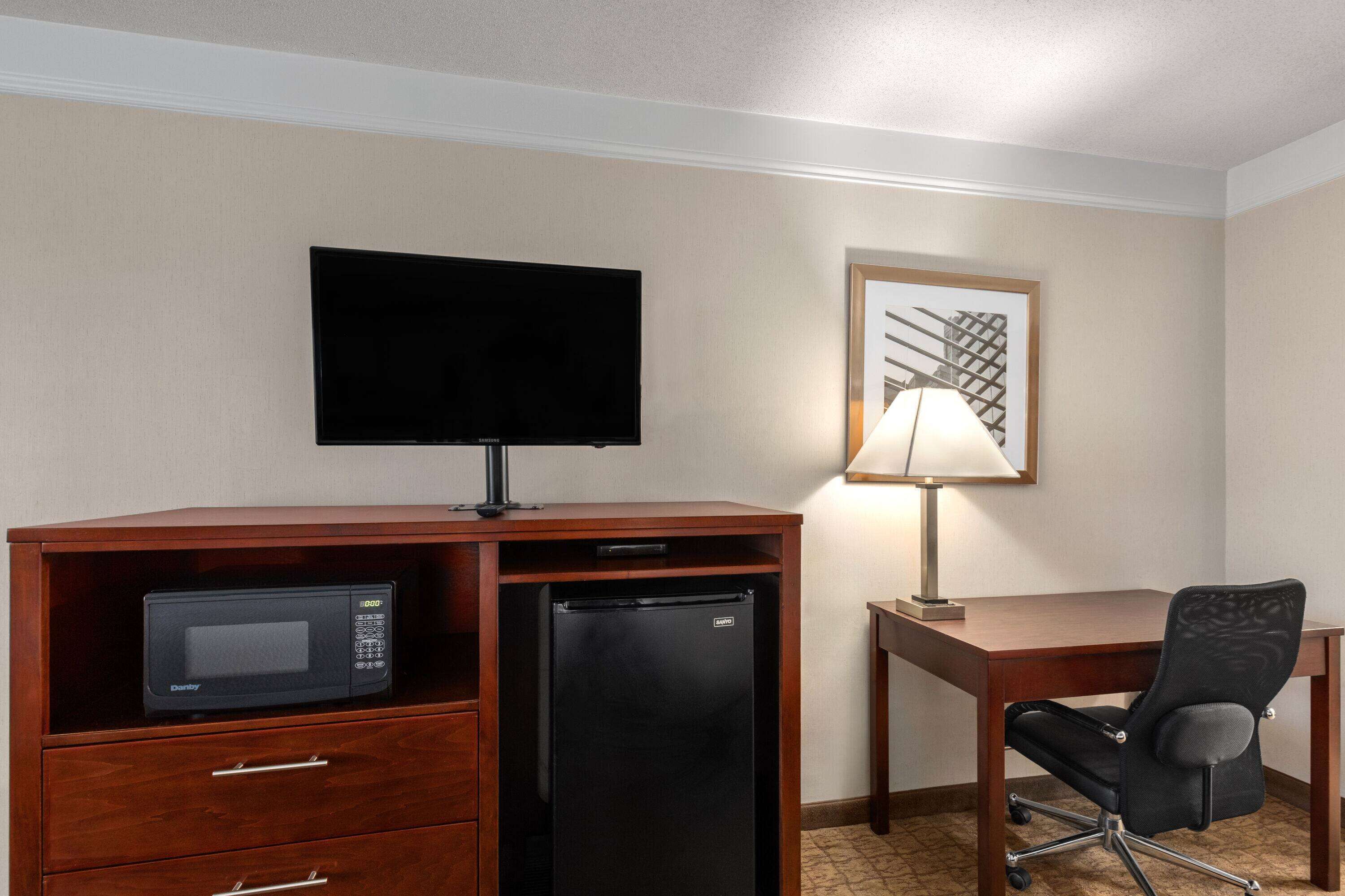 A hotel room with a wooden desk, a black refrigerator, a microwave, a flat-screen TV, a desk lamp, and a black office chair.