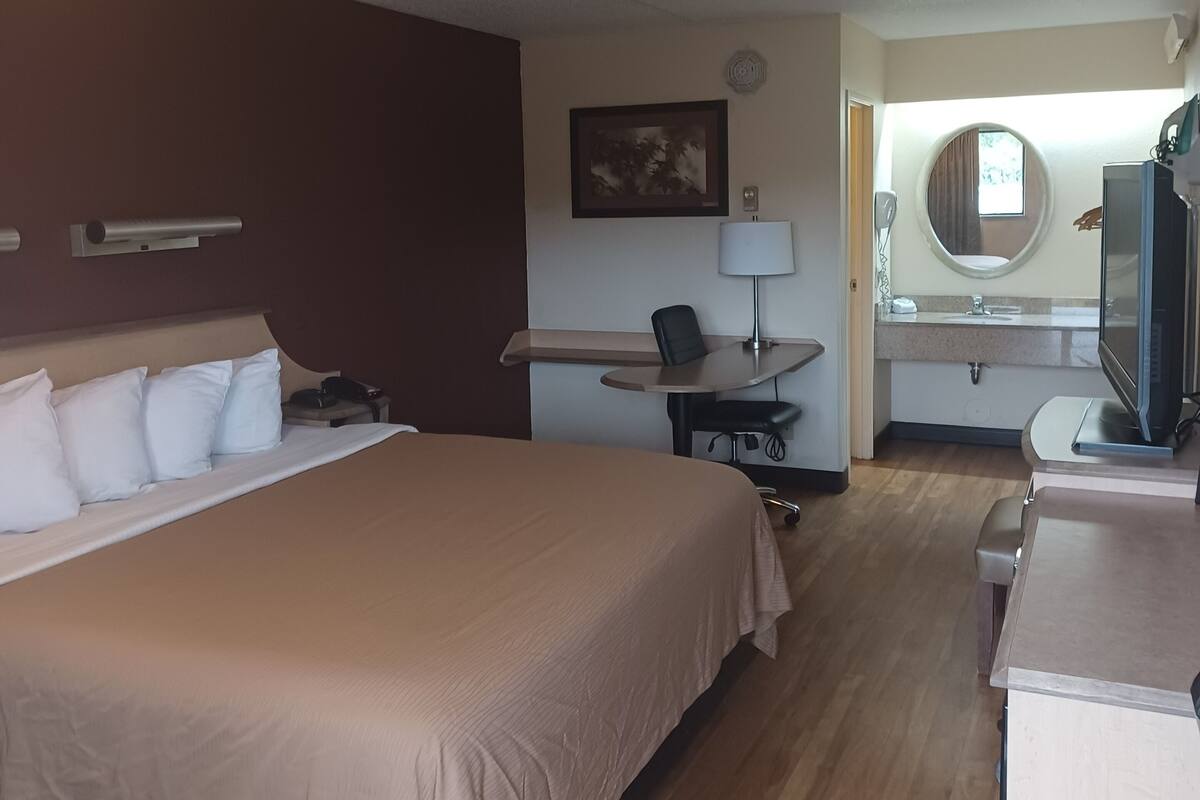 A hotel room with a large bed, a desk with a chair, a television, a round mirror, and a window.