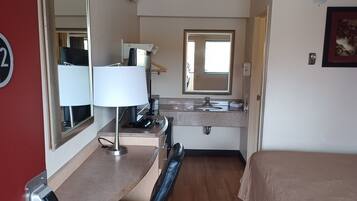 A hotel room with a bed, a desk with a chair, a mirror, a window, and a bathroom with a sink and toilet.