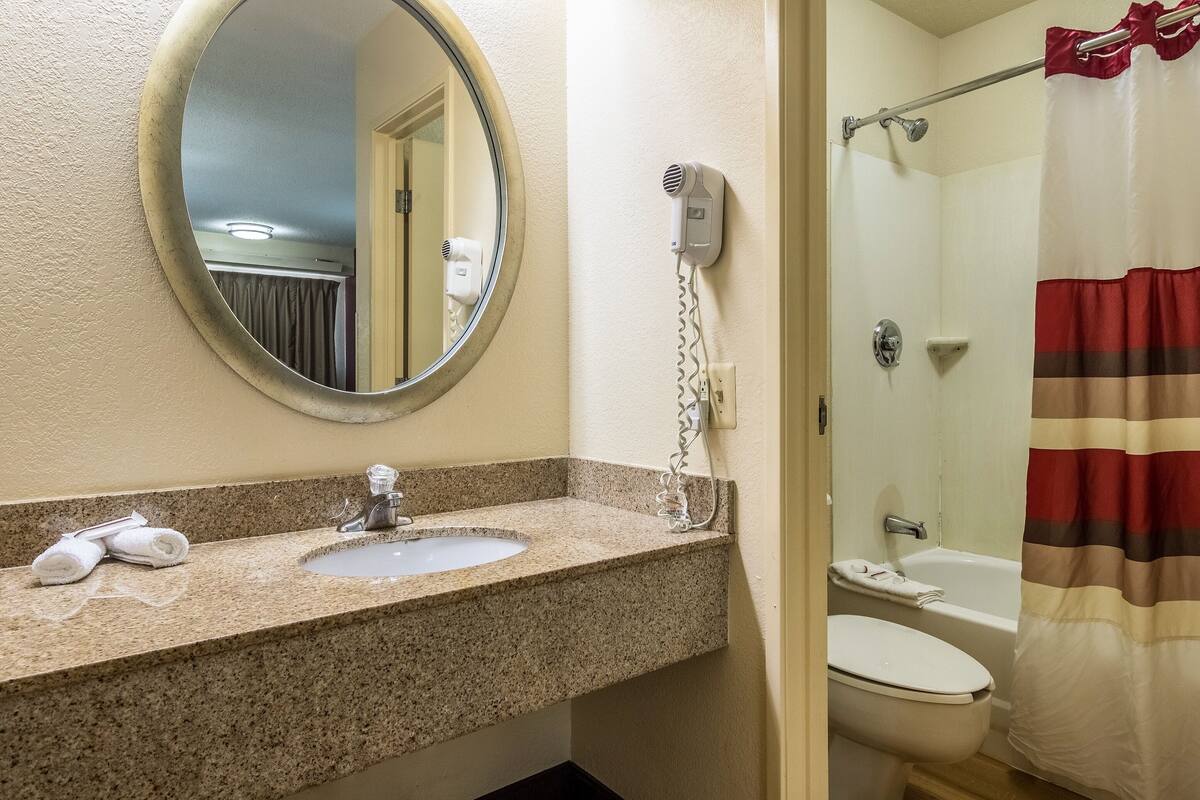A bathroom with a granite countertop sink, a round mirror, a hairdryer, a shower curtain, and a toilet.