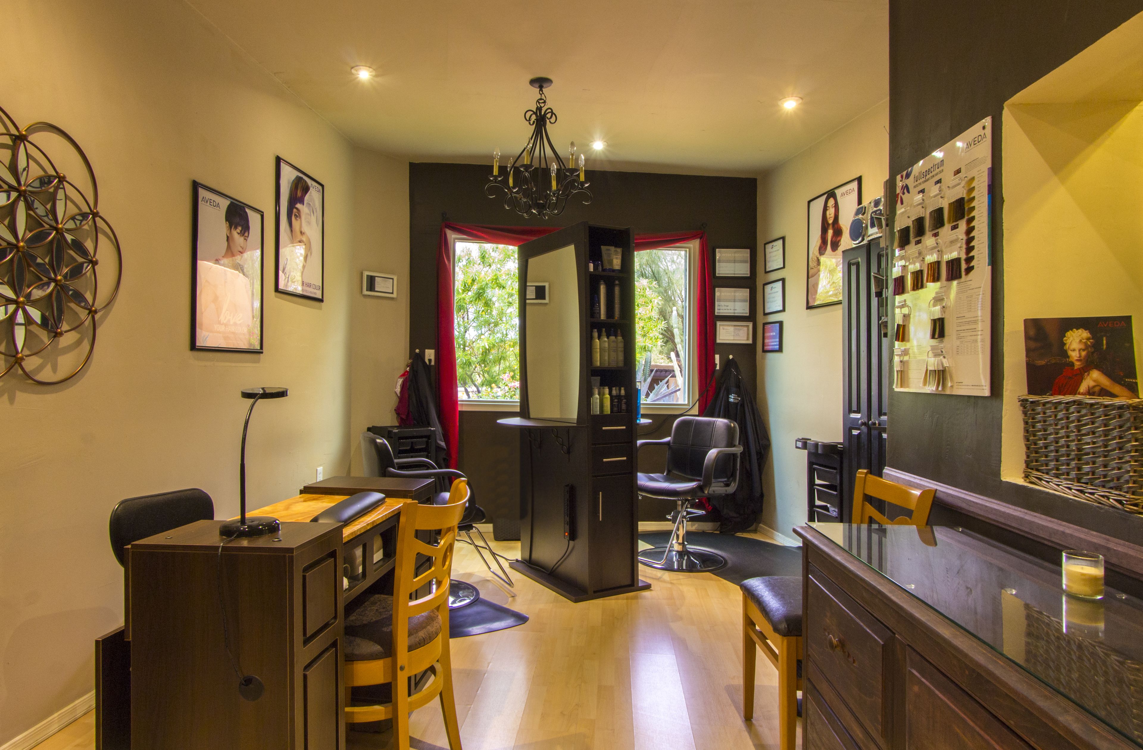 A well-lit room with a desk, chairs, a mirror, and a chandelier. There are framed pictures on the walls and a window with red curtains.