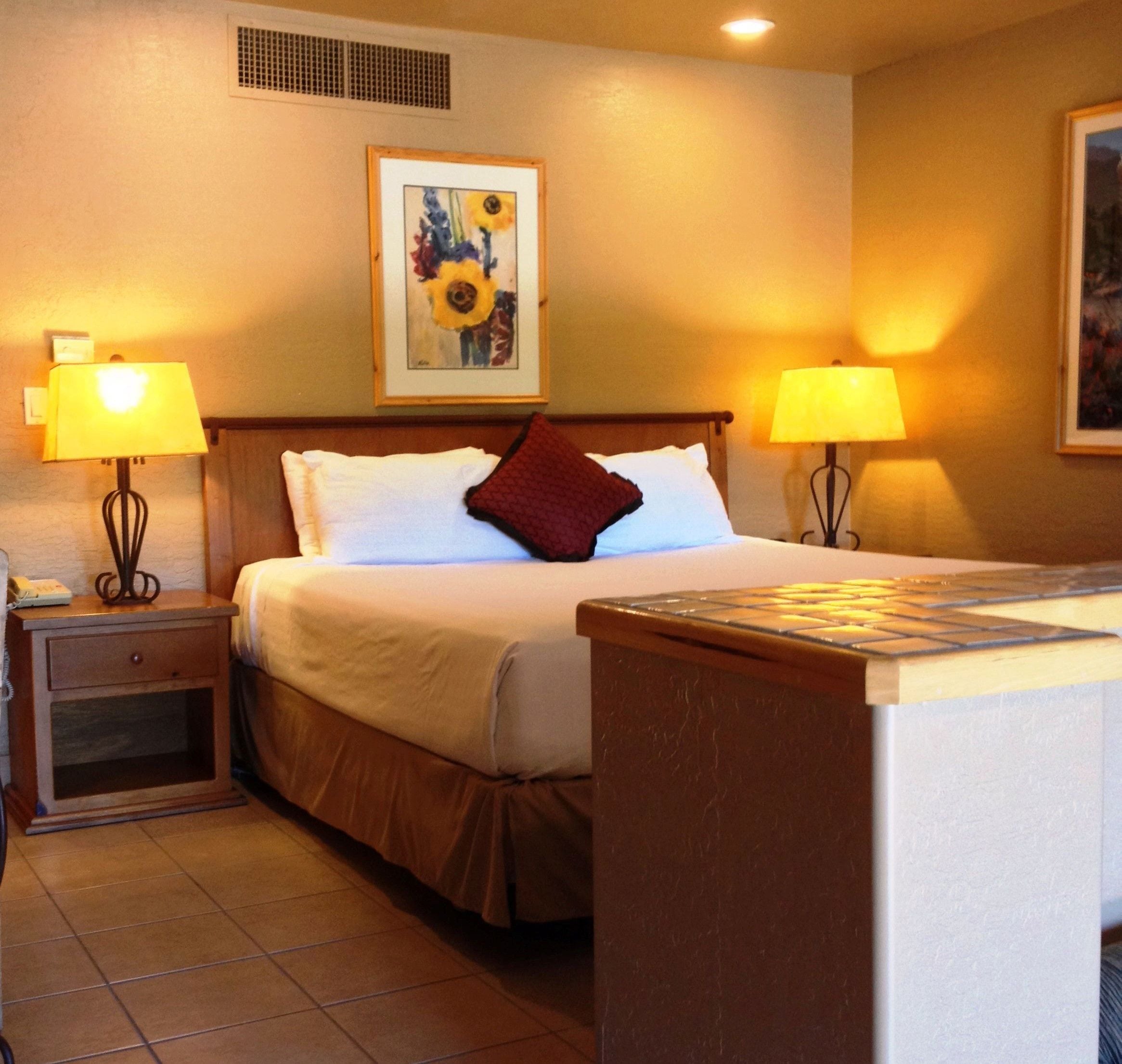 Una habitación de hotel con una cama, dos lámparas de noche, una mesita de noche, un cuadro de girasoles y suelo de baldosas.