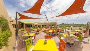 Une terrasse extérieure avec des tables et des chaises jaunes et orange, entourée de toiles d’ombrage orange.