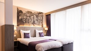 Una habitación de hotel moderna con una cama grande, un cuadro de un paisaje urbano en la pared y un ventanal con cortinas.