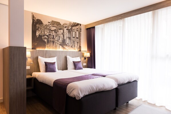A modern hotel room with a large bed, a cityscape artwork on the wall, and a large window with curtains.