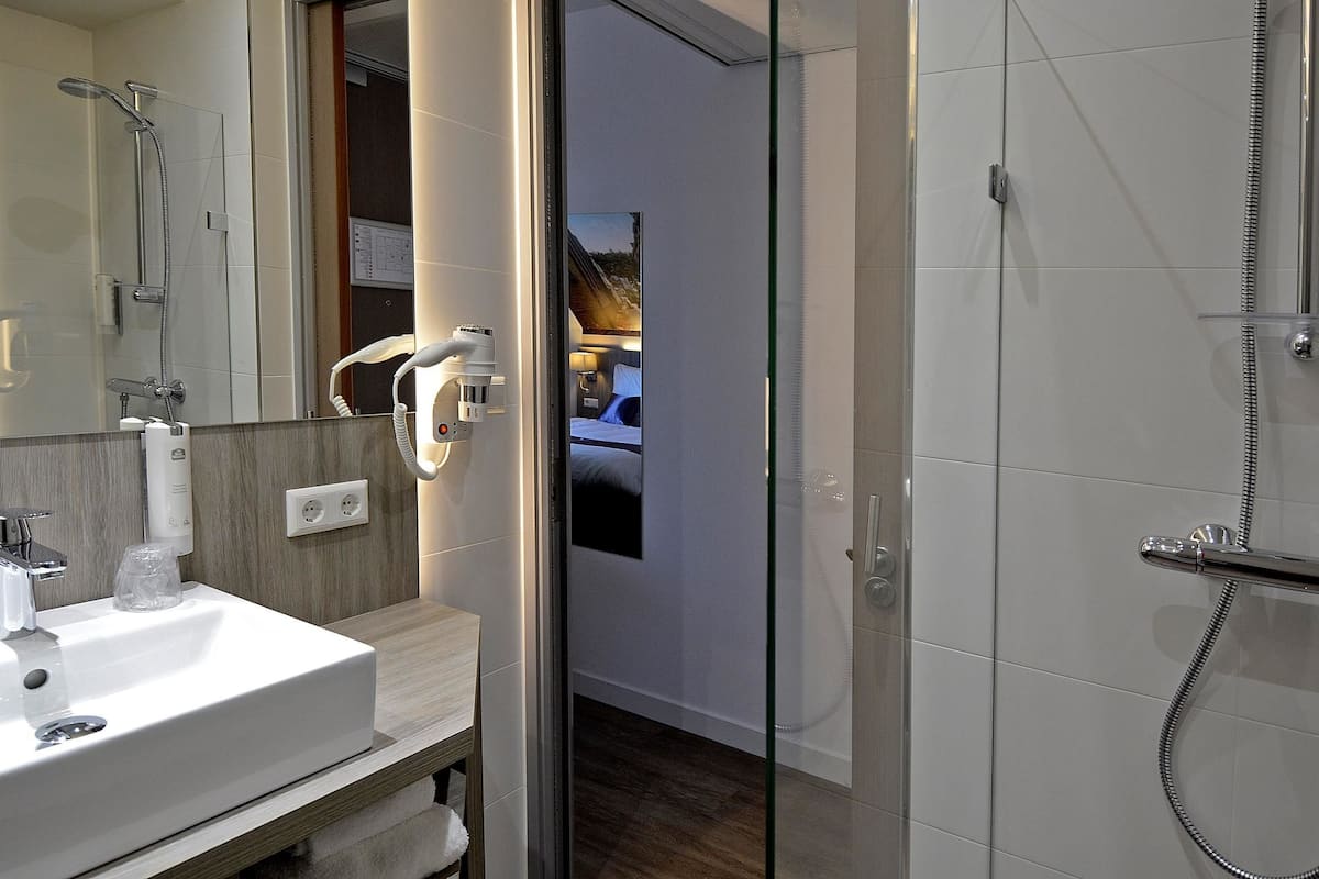 Une salle de bain moderne avec un lavabo blanc, une cabine de douche en verre et une chambre à coucher visible à l’arrière-plan.