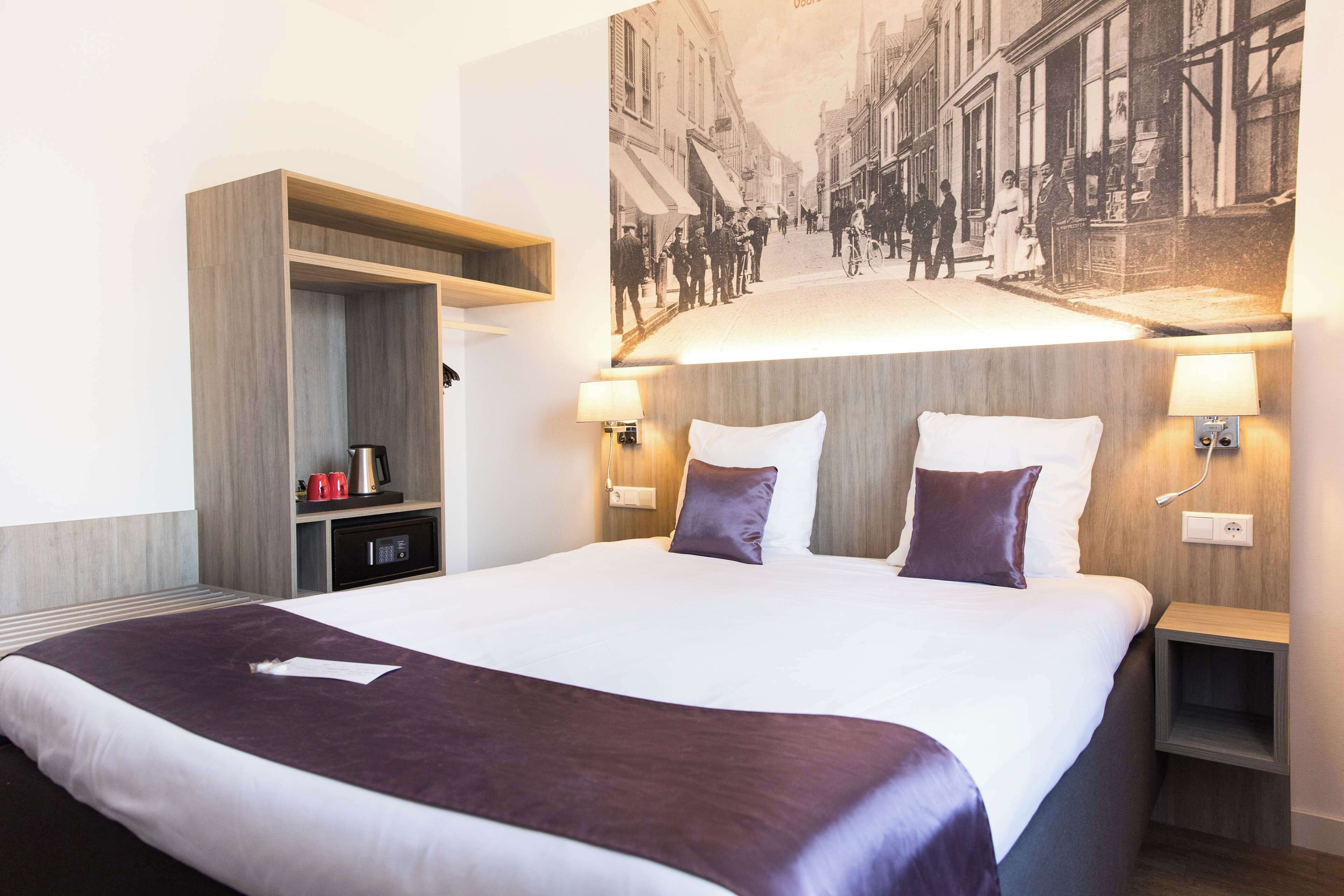 A hotel room with a large bed, a built-in wardrobe, bedside lamps, and a vintage street scene on the wall.