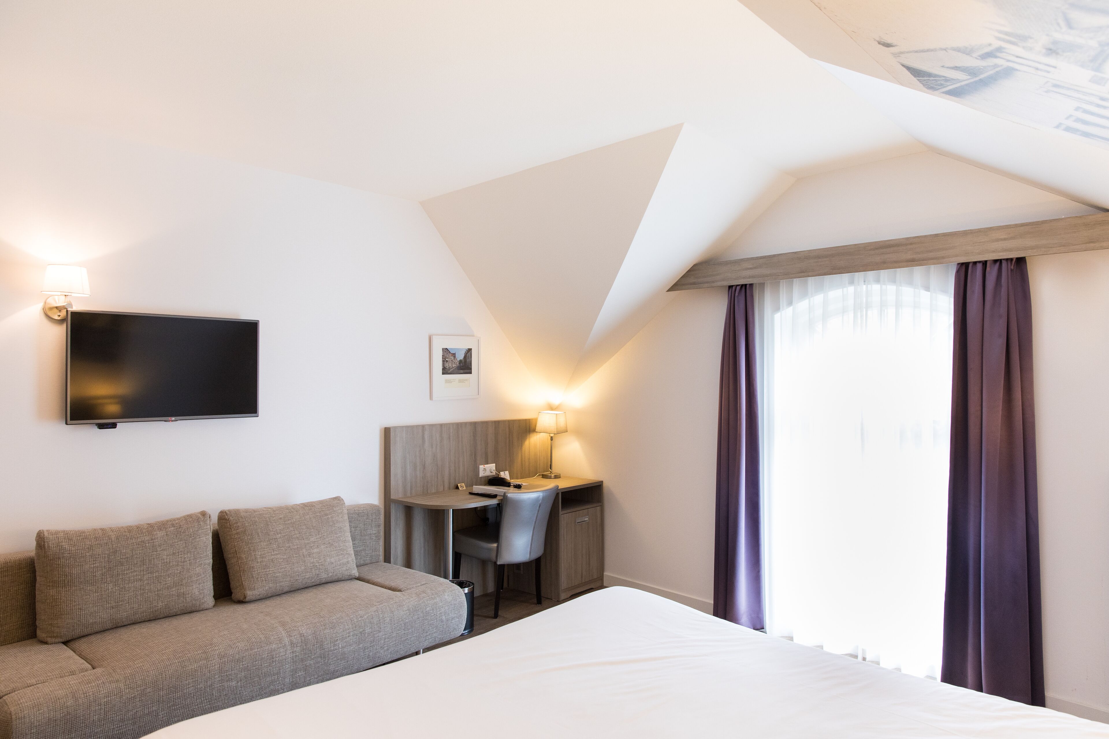 A hotel room with a bed, a desk, a chair, a flat-screen TV, and a window with curtains.