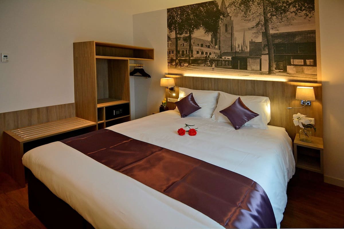 Une chambre d’hôtel avec un grand lit, une armoire en bois, une table de chevet avec une lampe et un tableau accroché au mur.