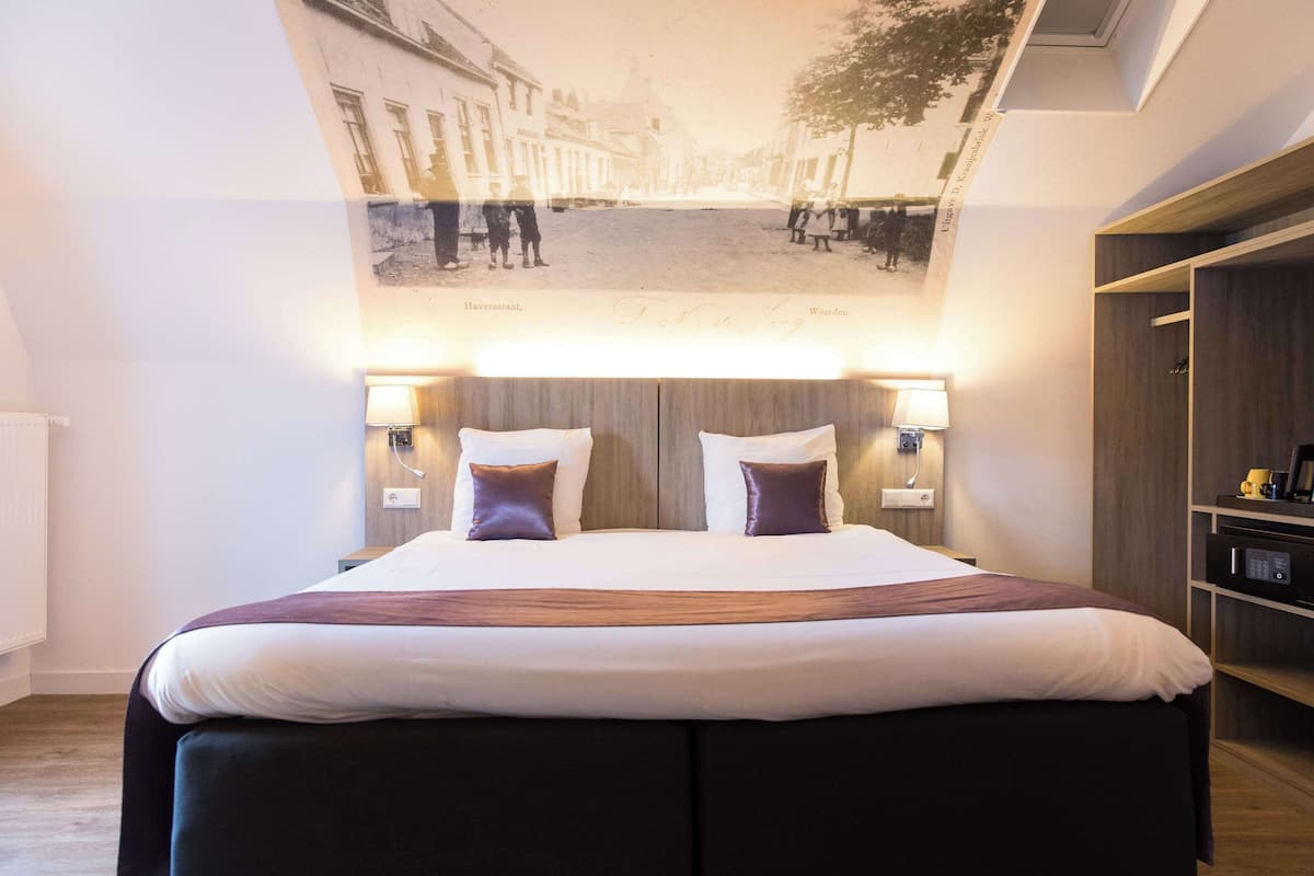 Une chambre d’hôtel moderne, dotée d’un grand lit, de deux lampes de chevet, d’une tête de lit en bois et d’une œuvre d’art murale représentant une scène de rue.