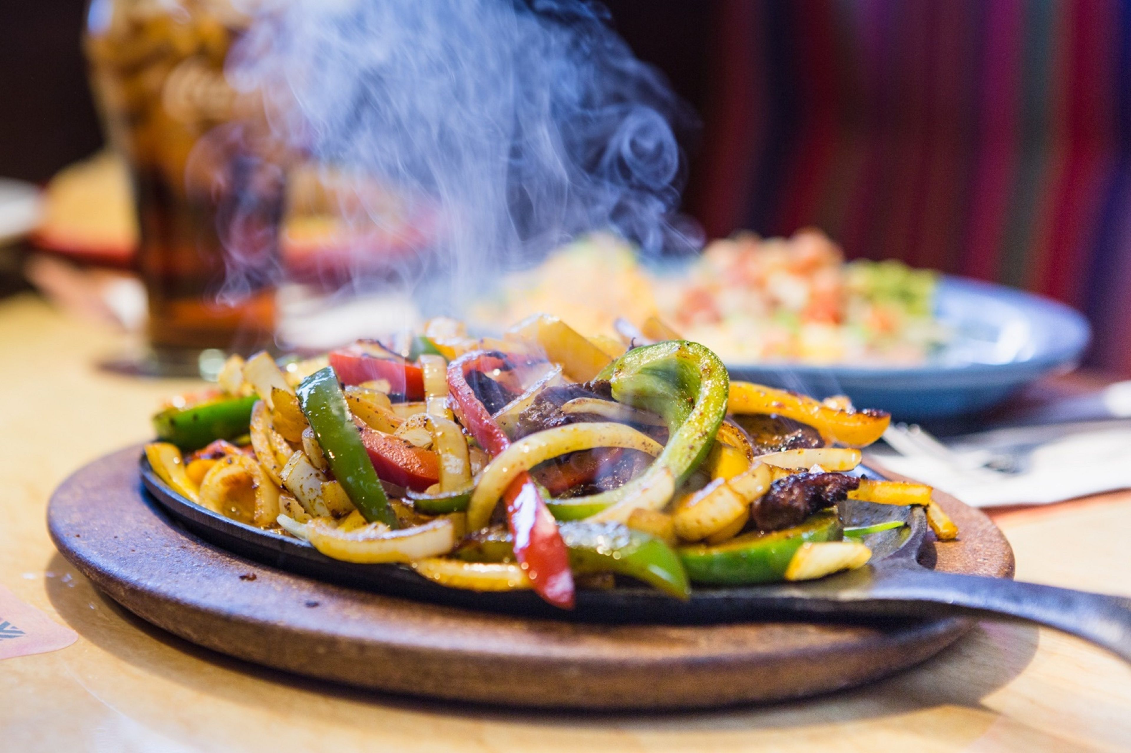 A close-up of a sizzling vegetable dish on a wooden board, steam rising from the food, suggesting a restaurant setting.