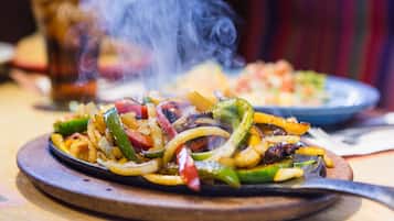 A close-up of a sizzling vegetable dish on a wooden board, steam rising from the food, suggesting a restaurant setting.