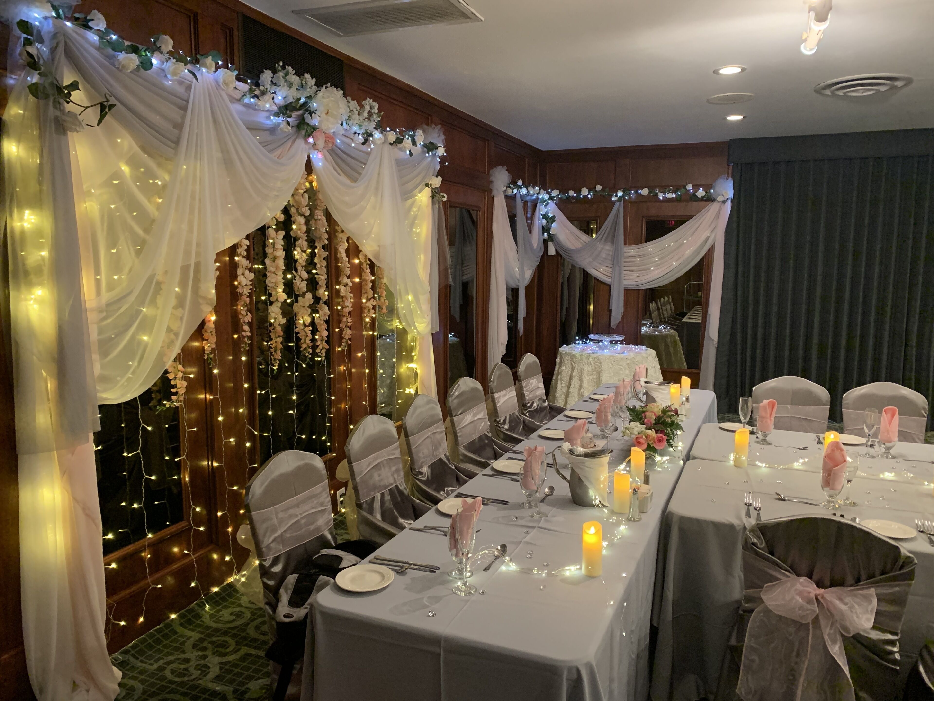 Um salão de banquetes com mesas montadas para um evento formal, coberto com tecido branco e decorações florais.