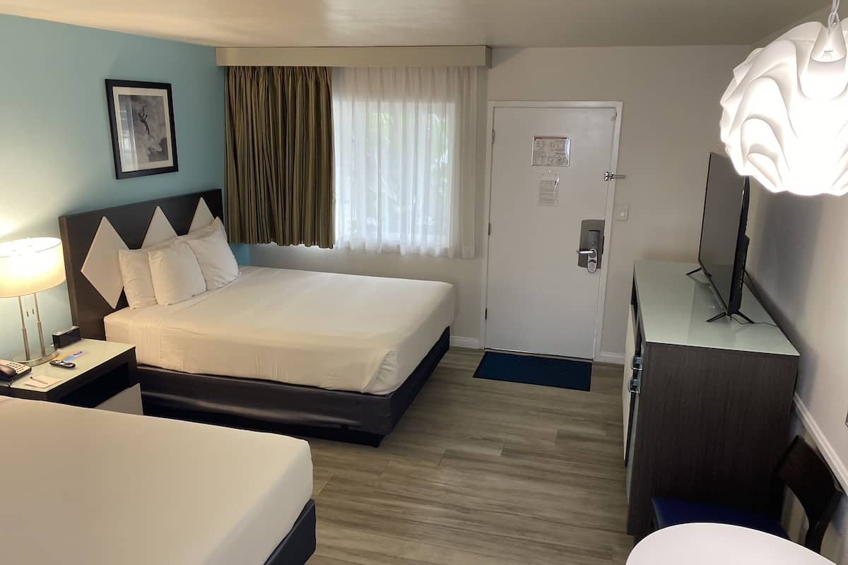 A hotel room with a bed, bedside table, lamp, TV, and a door with a security panel.