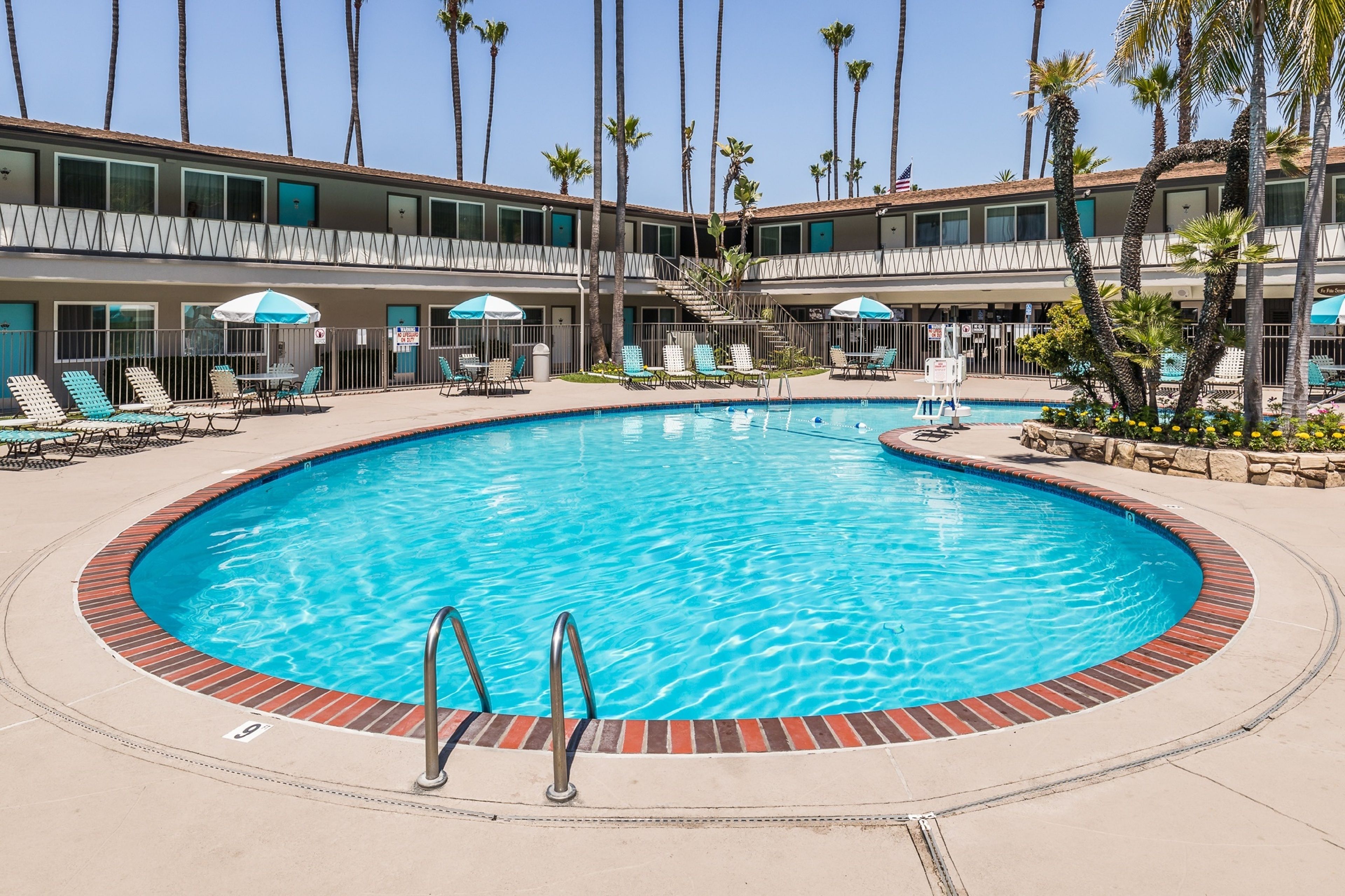 Une piscine entourée de chaises longues et de parasols, bordée de palmiers et d’un bâtiment d’hôtel.