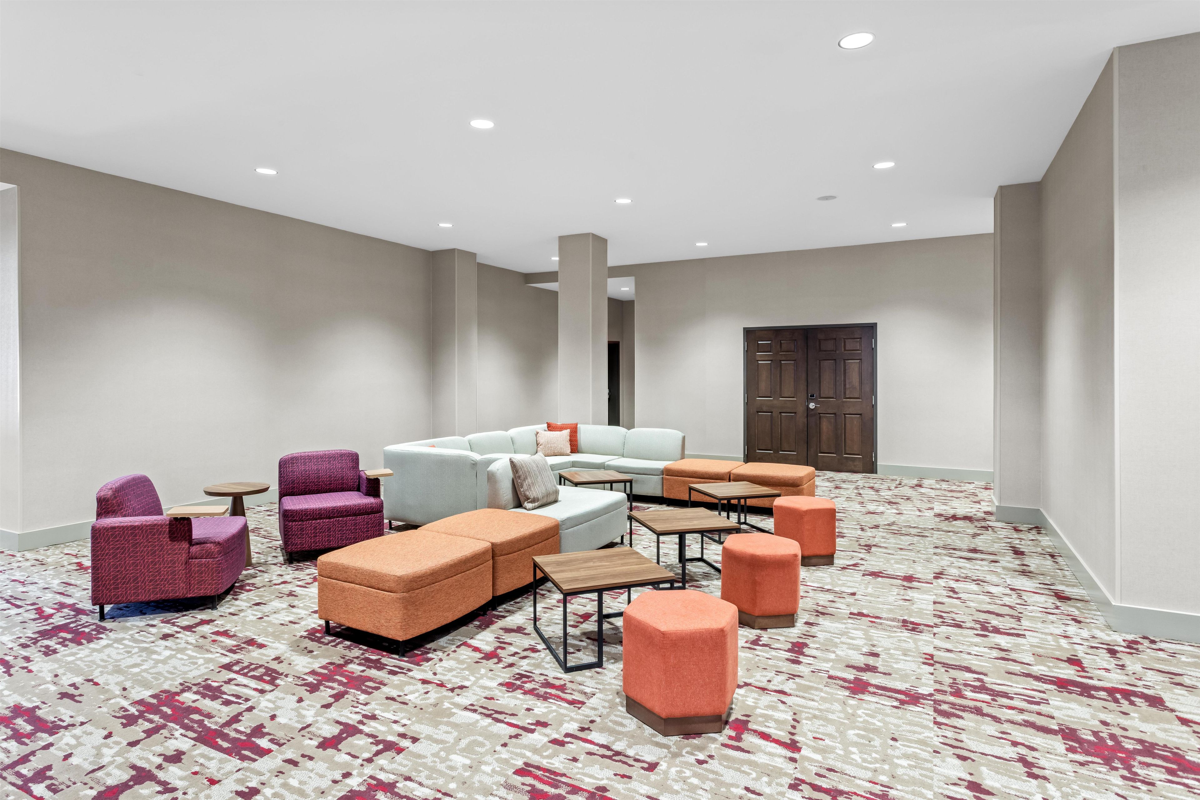 Un hall d’hôtel doté d’un espace détente comprenant des canapés, des fauteuils et des poufs, ainsi qu’une moquette à motifs et une porte en bois.