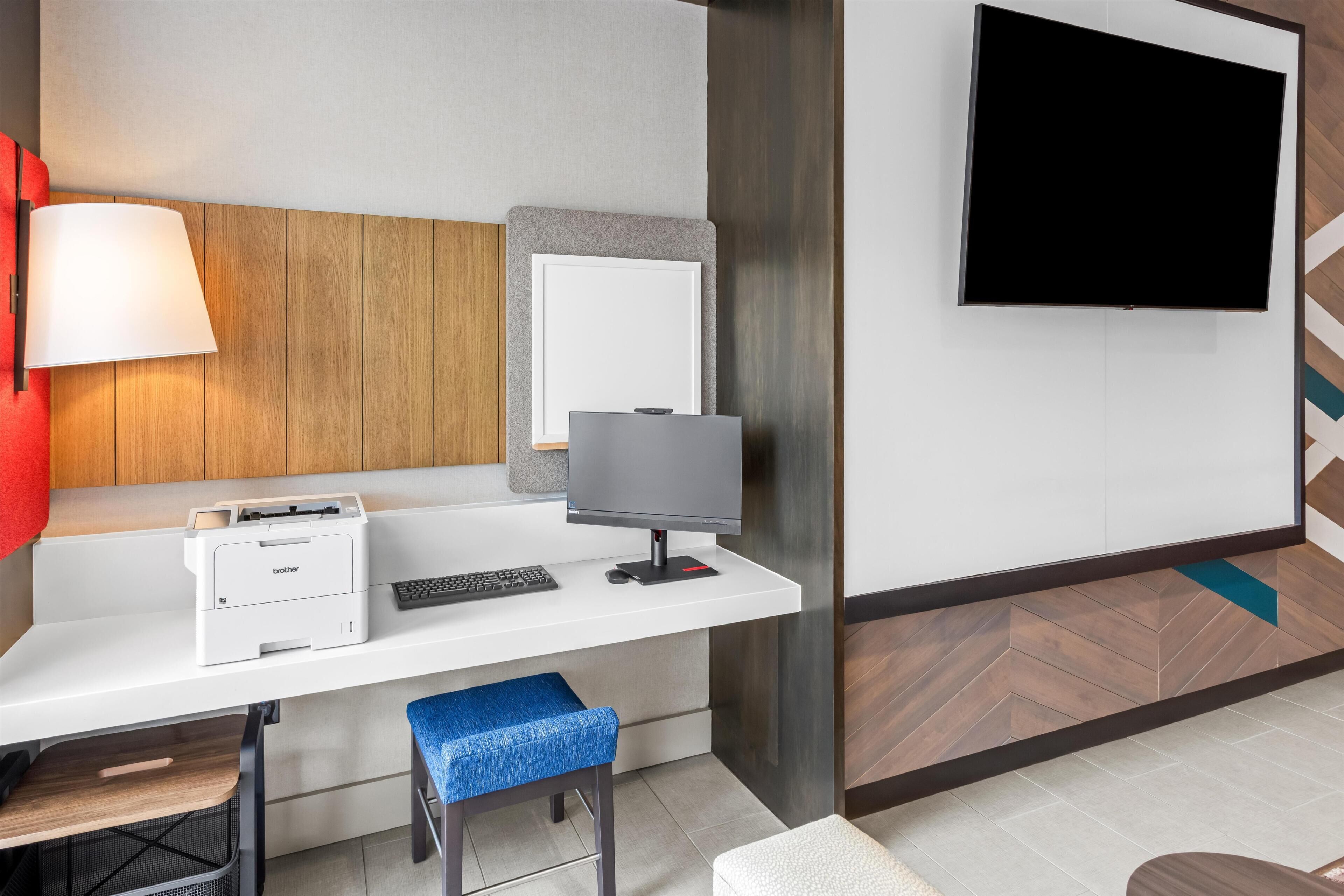 A hotel room with a desk, a flat-screen TV, a lamp, a printer, and a chair.
