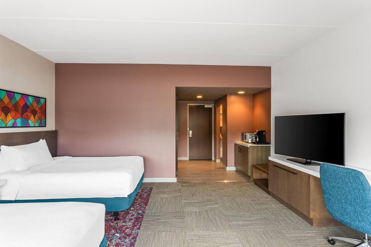 Ein Hotelzimmer mit zwei Betten, einem Fernseher, einem Schreibtisch und einer Küchenzeile.