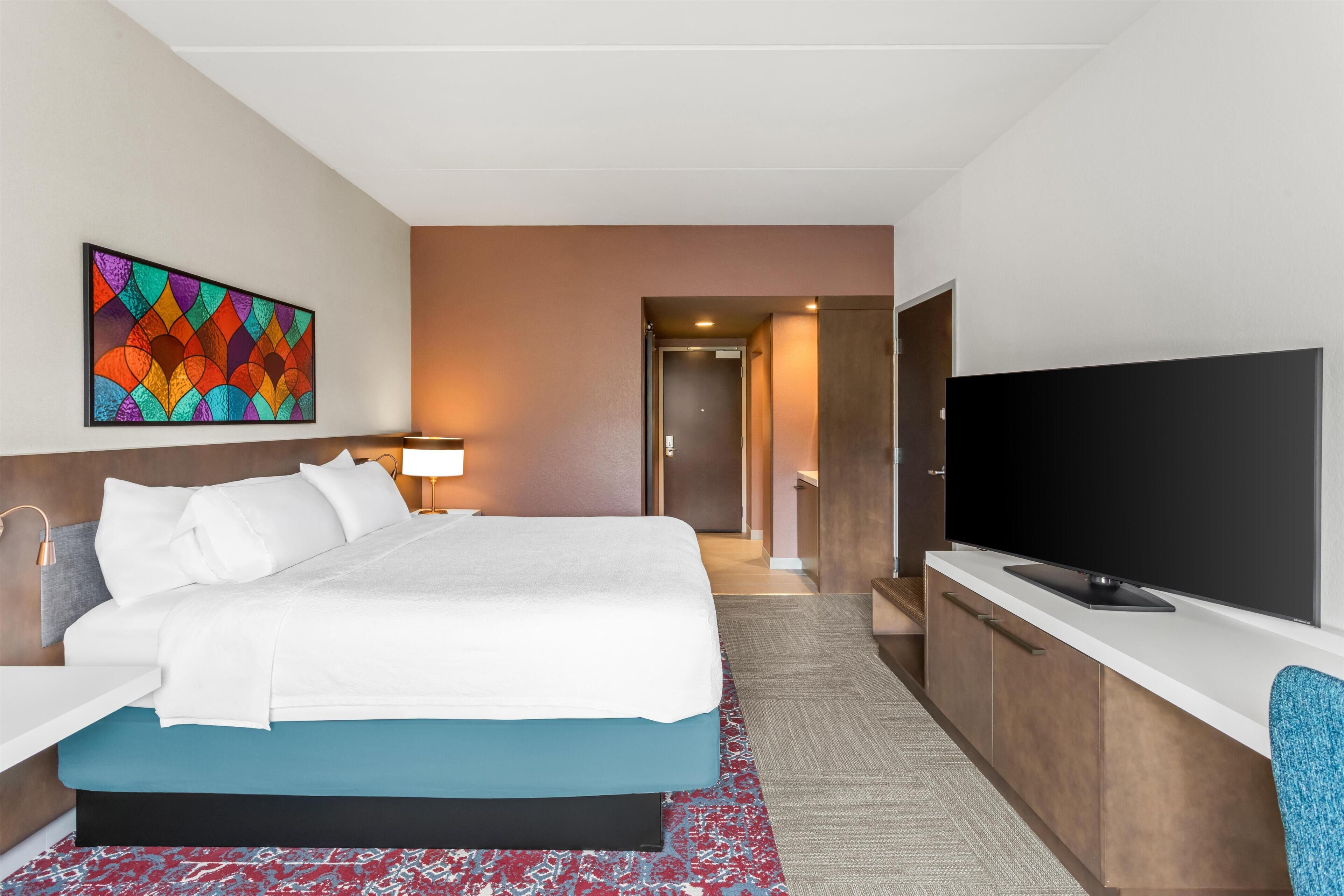 A hotel room with a bed, a TV, a desk, and a colorful artwork on the wall.