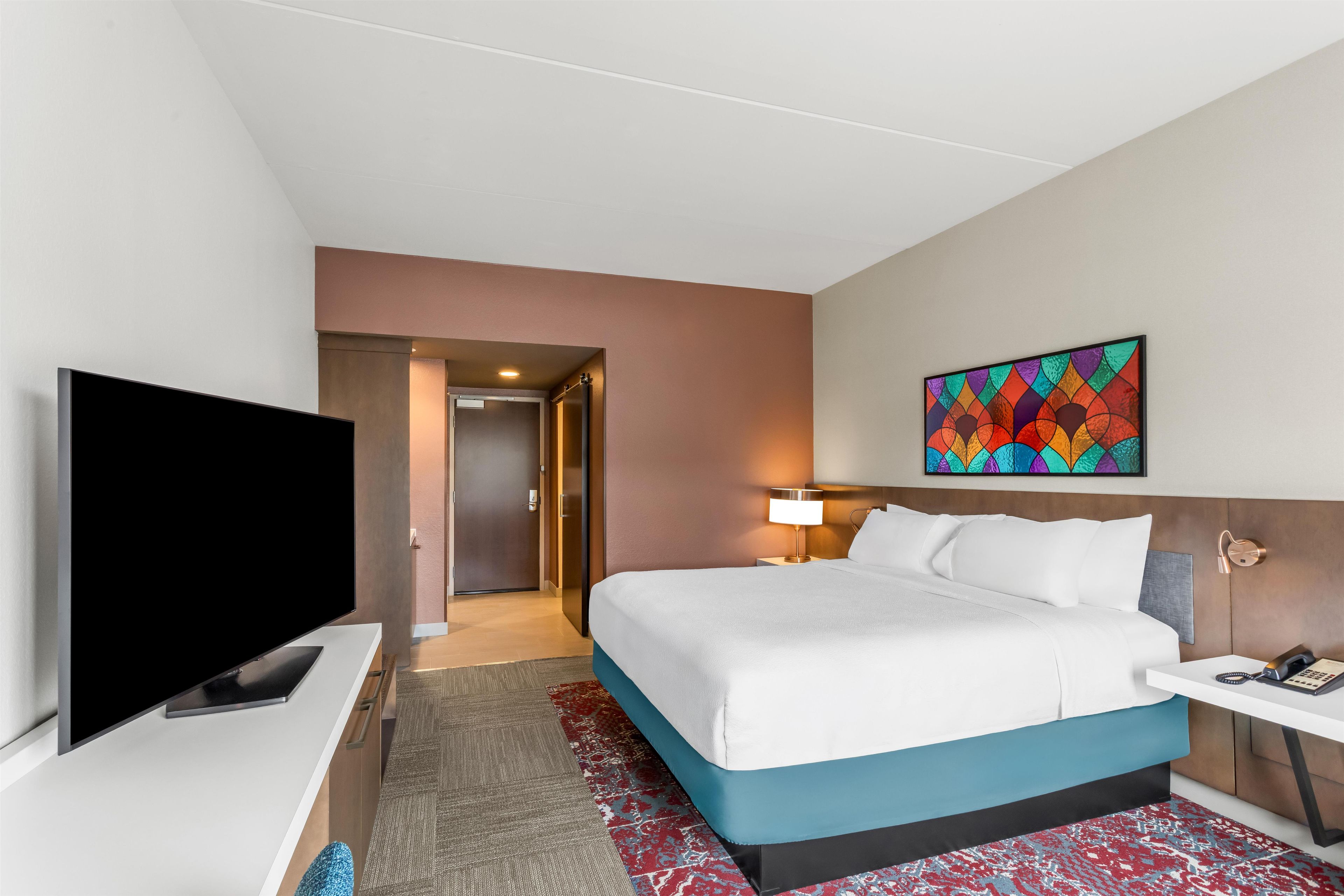 A hotel room with a bed, a TV, a desk, a lamp, and a painting on the wall.