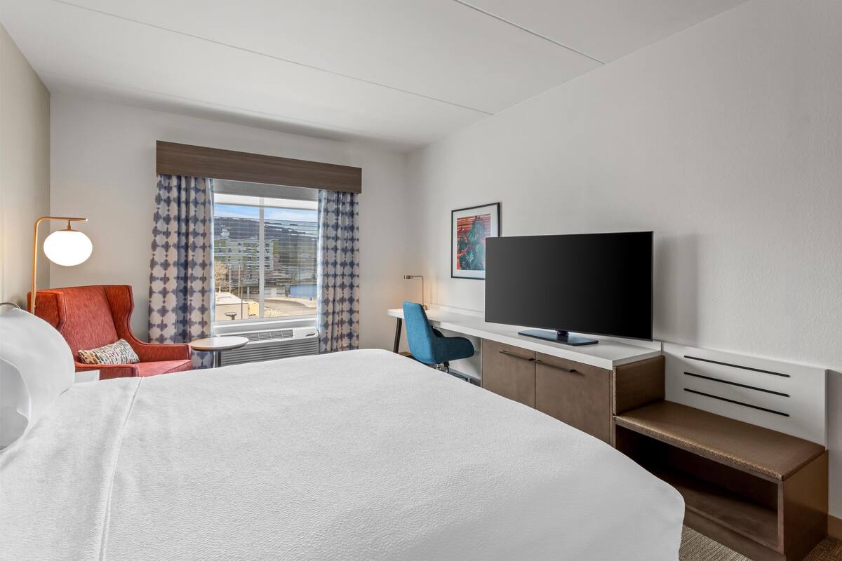 Ein Hotelzimmer mit einem großen Bett, einem Schreibtisch mit Fernseher, einem Sessel, einer Lampe und einem Fenster mit Vorhängen.