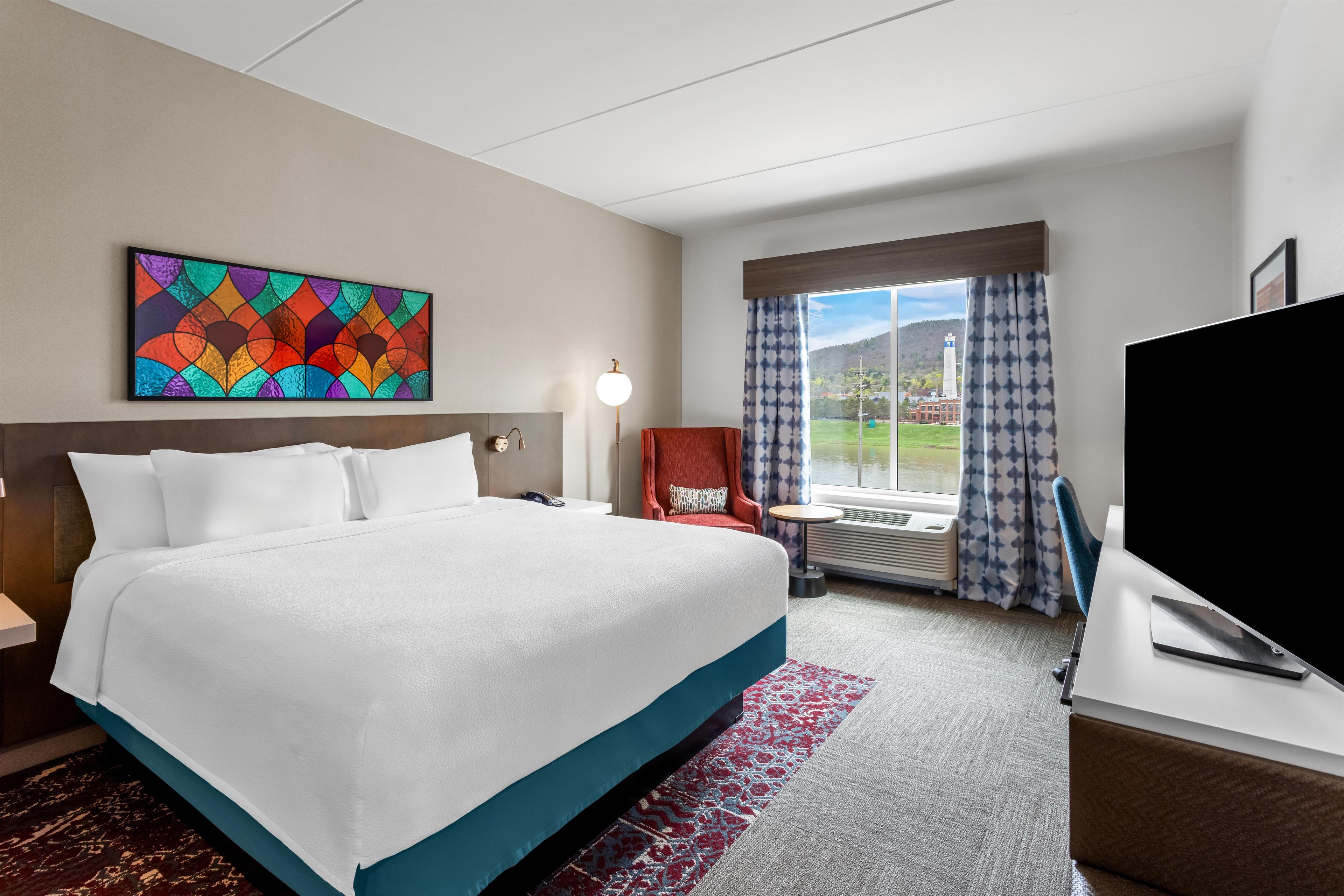 Ein Hotelzimmer mit einem großen Bett, einem Schreibtisch mit Flachbildfernseher, einem Sessel, einer Schreibtischlampe und einem Fenster mit Blick auf ein Gebäude und Grünflächen.