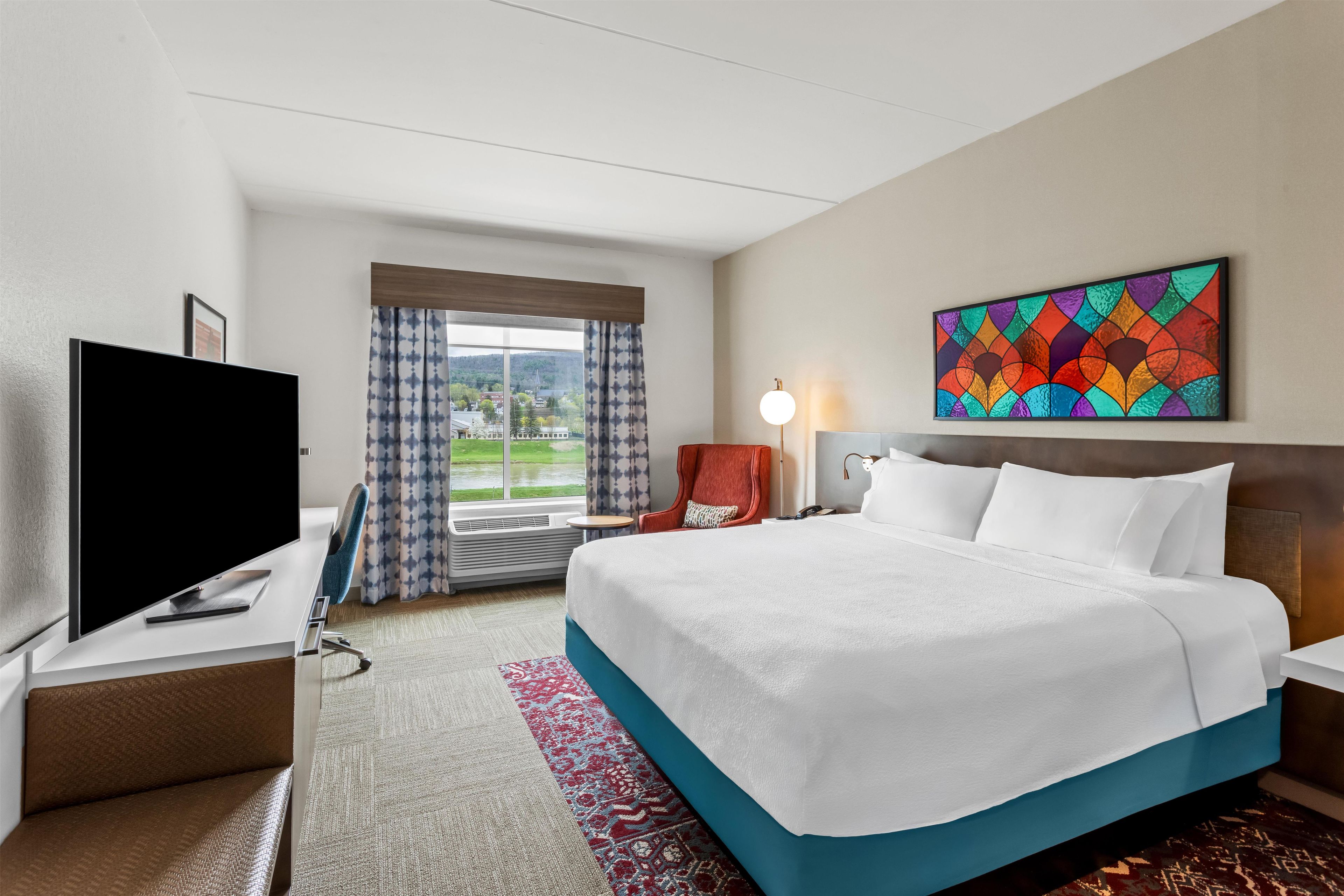 Quarto de hotel com cama, mesa de trabalho com televisão, cadeira, mesa de cabeceira, abajur e janela com cortinas.