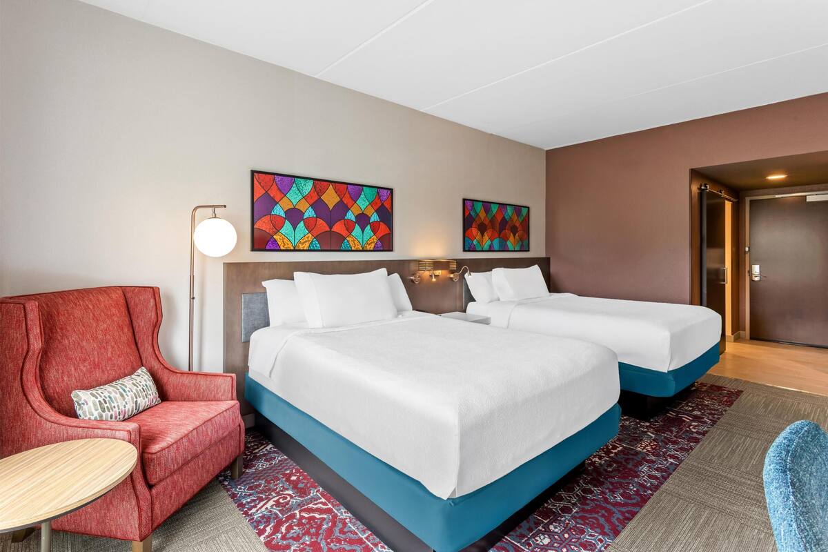 Ein Hotelzimmer mit zwei Betten, einem roten Sessel, einem kleinen runden Tisch und zwei farbenfrohen Wandbildern.