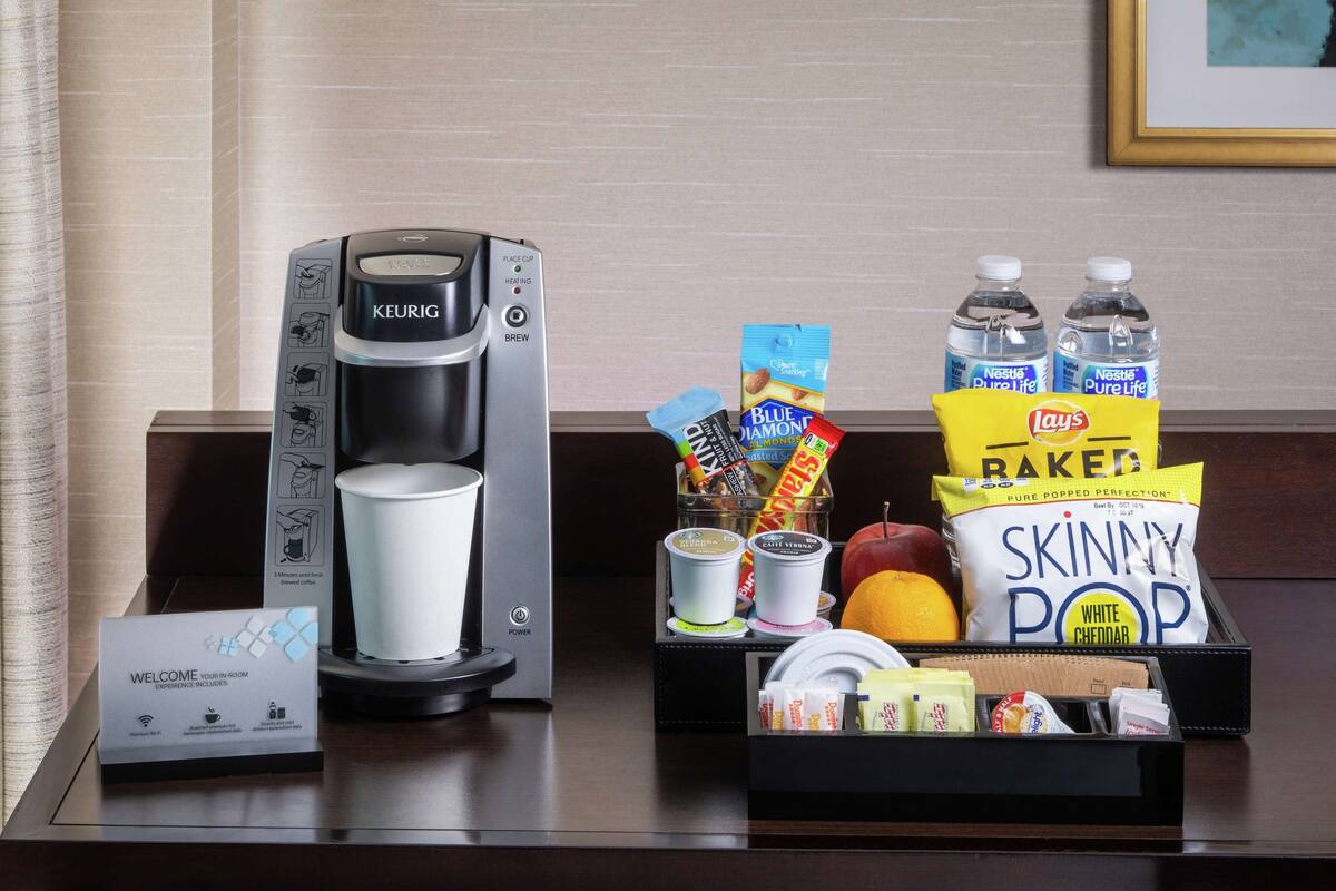 Eine Kaffeemaschine, eine Willkommenskarte, Snacks und Wasserflaschen auf einem Hotel-Schreibtisch.