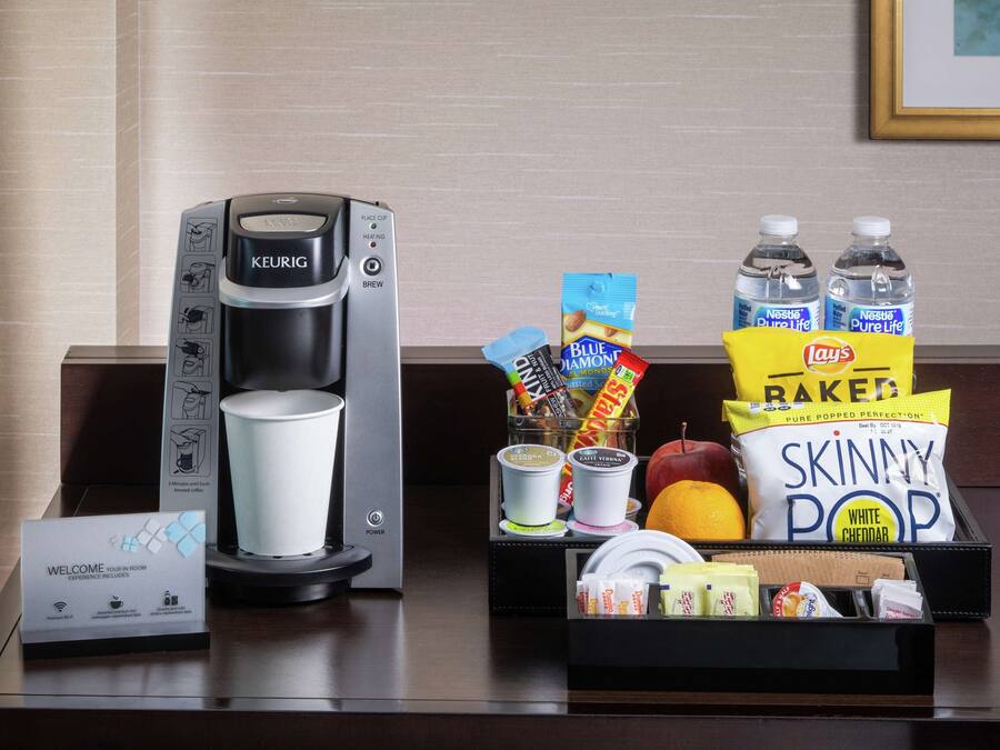 Una cafetera, una tarjeta de bienvenida, aperitivos y botellas de agua sobre un escritorio de hotel.